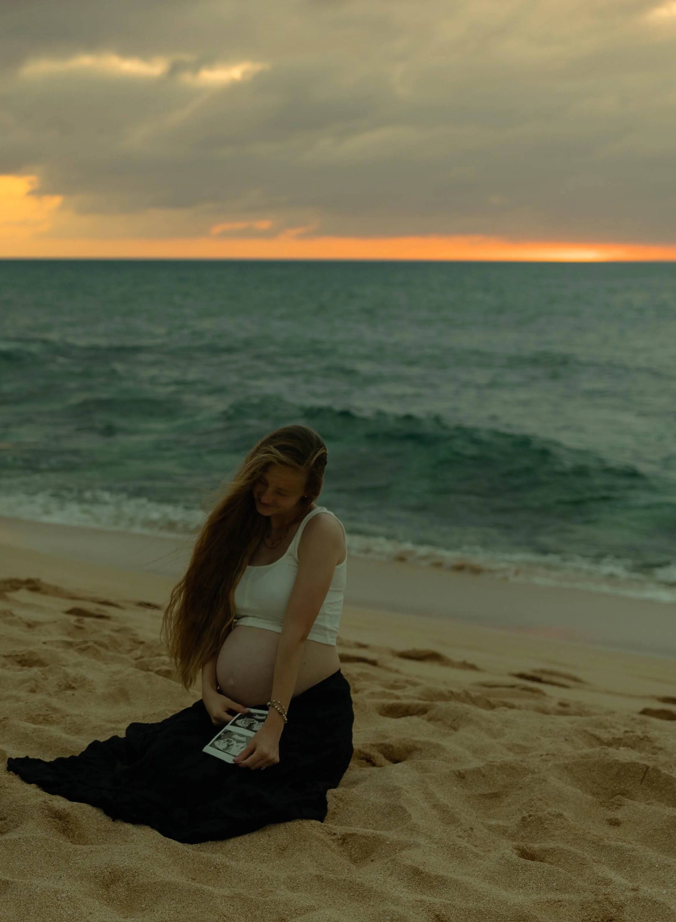 A woman with long hair sits on a sandy beach during sunset, looking at a photo album, with the ocean and cloudy sky in the background.