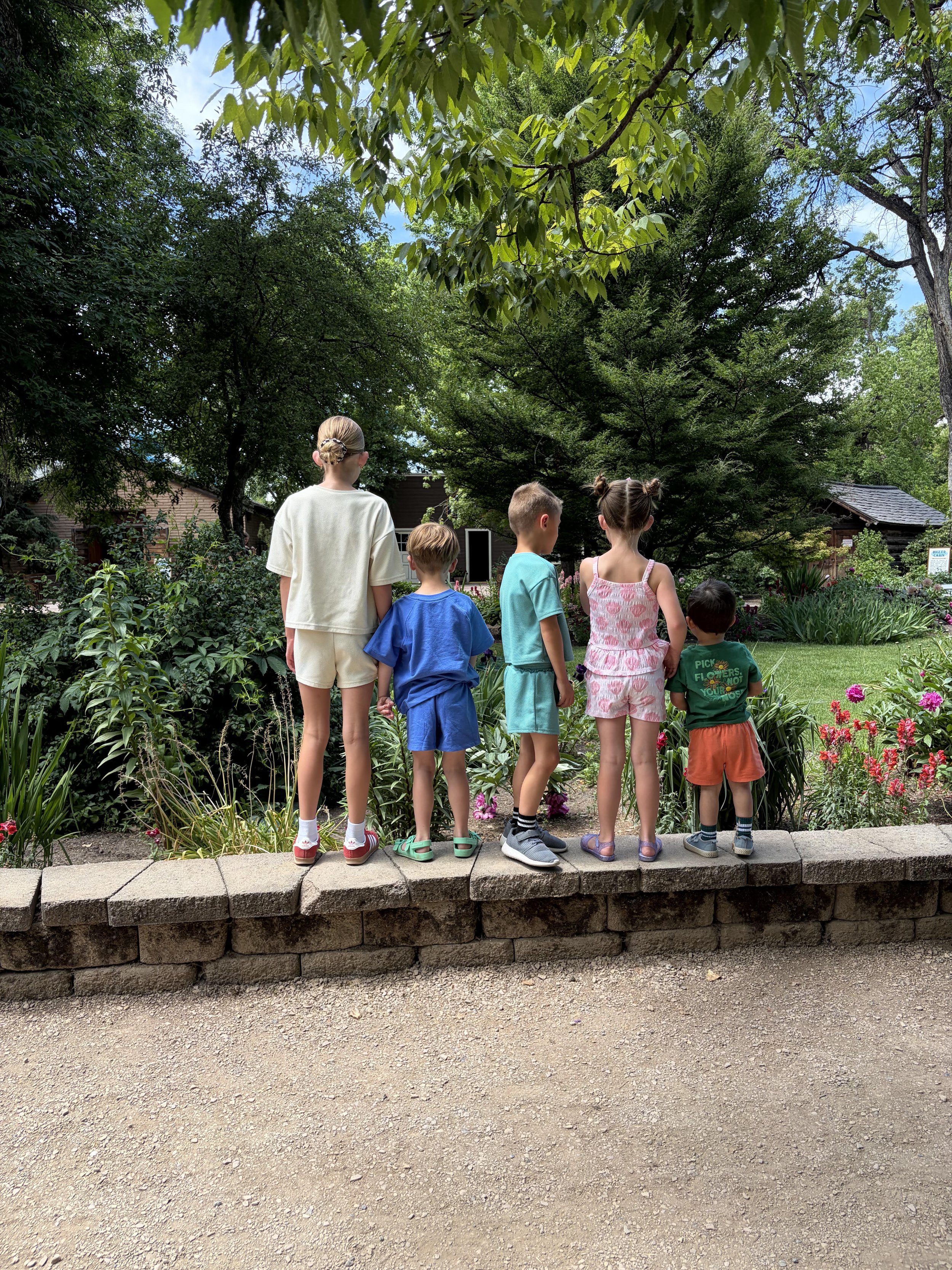 A group of five children standing on a brick ledge in a park or garden, facing away from the camera, with lush green trees and plants in the background.