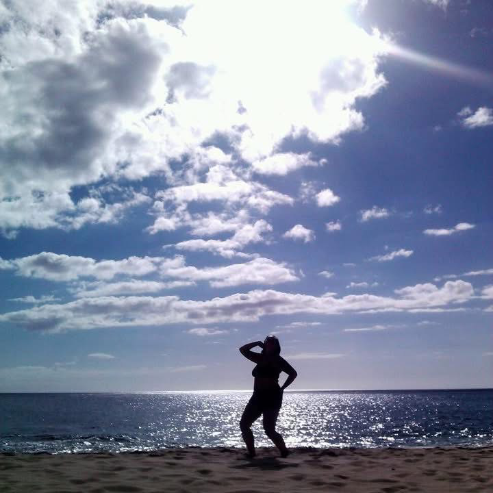 Silhouette of a person standing on a beach with one arm raised, facing the ocean under a partly cloudy sky with the sun shining.