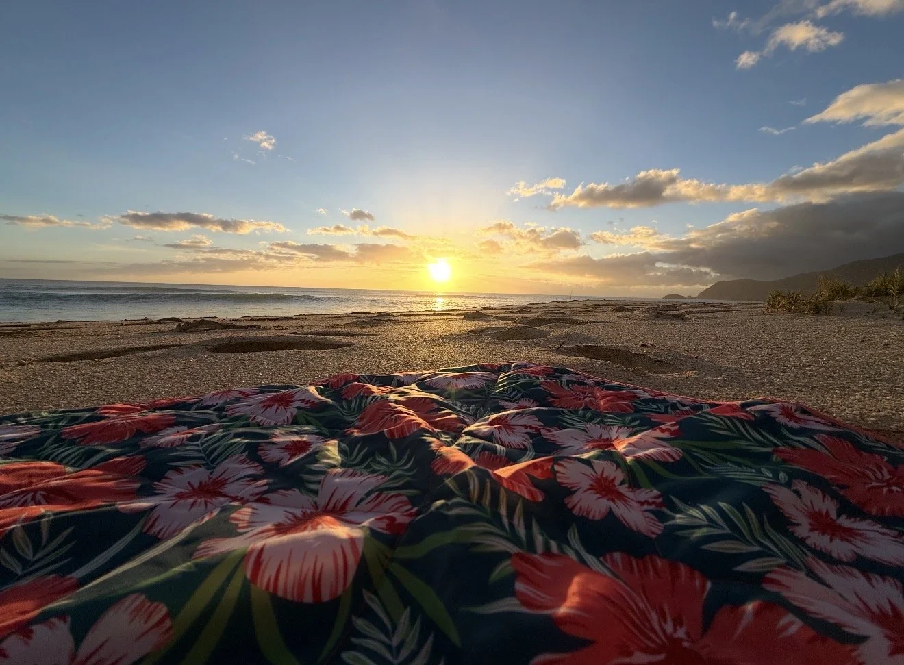 Beach at sunset with a floral blanket in the foreground and mountains in the background.