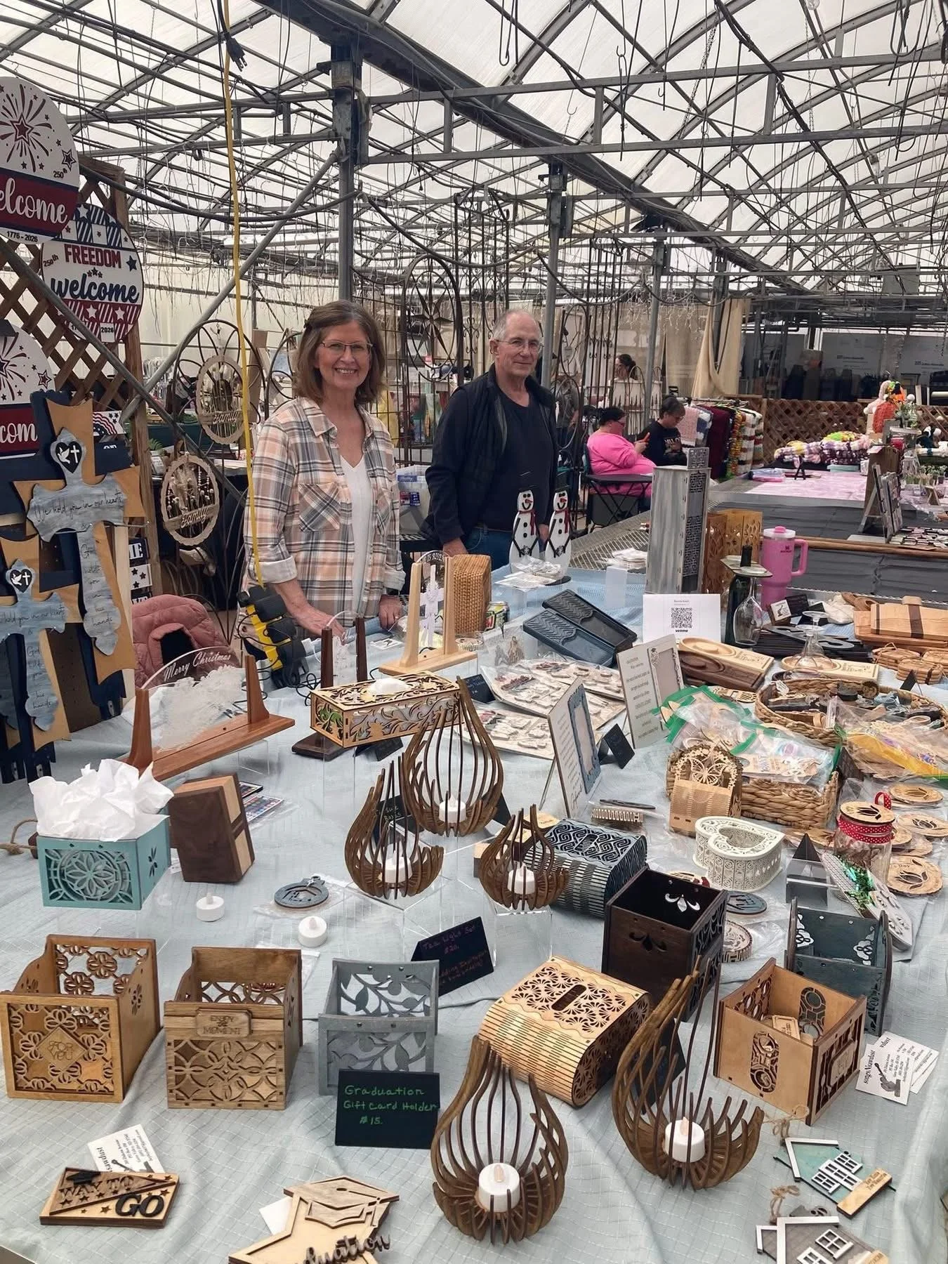 Two people standing behind a table displaying wooden crafts at a market stall inside a greenhouse or large indoor space.