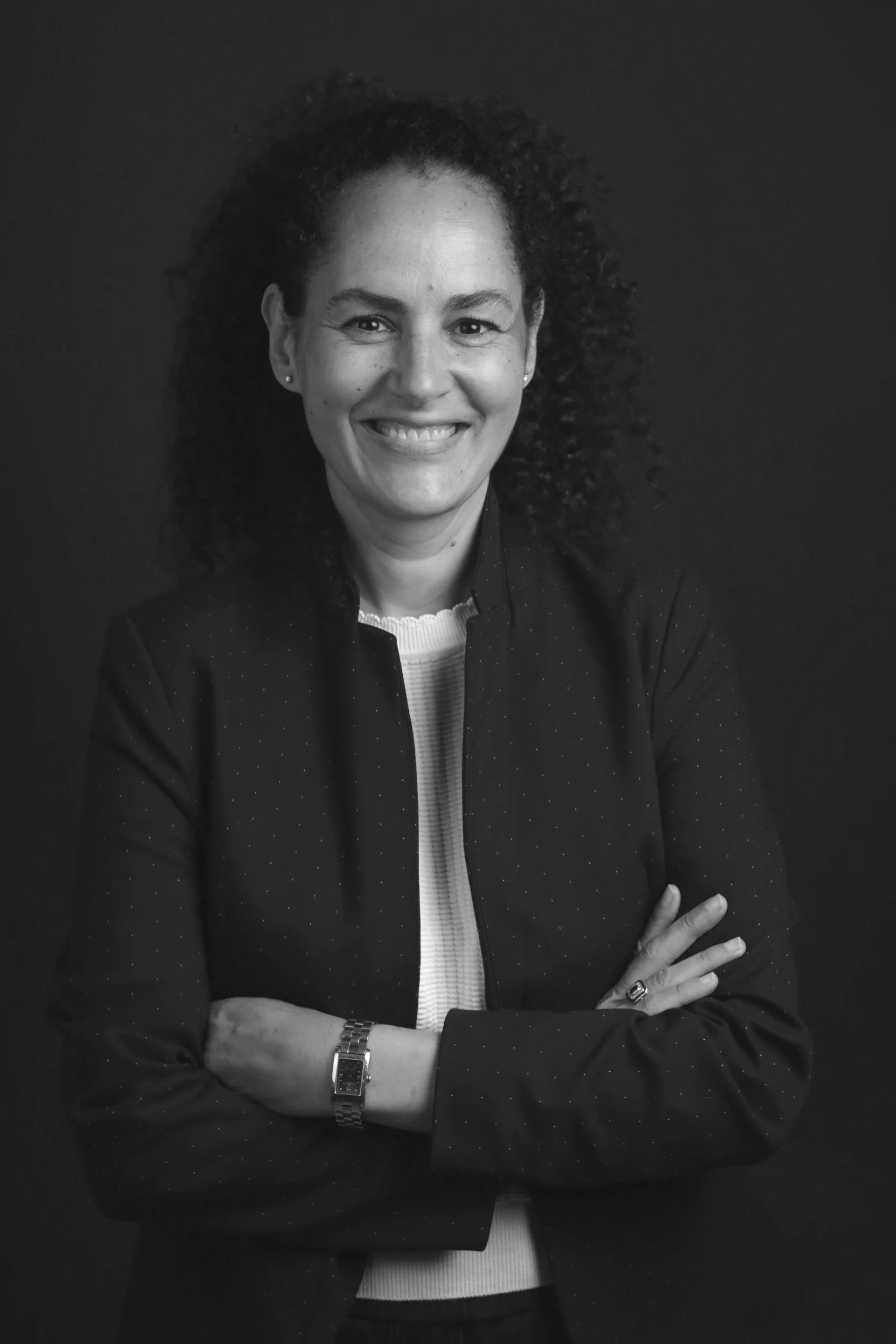 Une femme souriante avec des cheveux bouclés, portant une chemise noire et une montre, posant avec les bras croisés contre un fond sombre. directeur gouvernance l'Etudiant