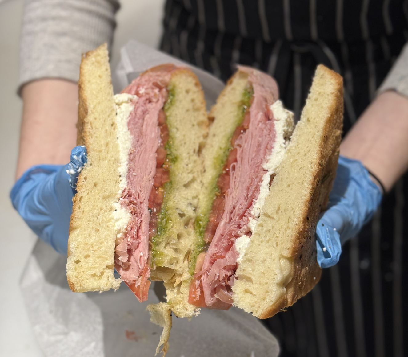 Close-up of a large sandwich with layers of deli meat, lettuce, tomato, and green sauce, held by a person wearing blue gloves.