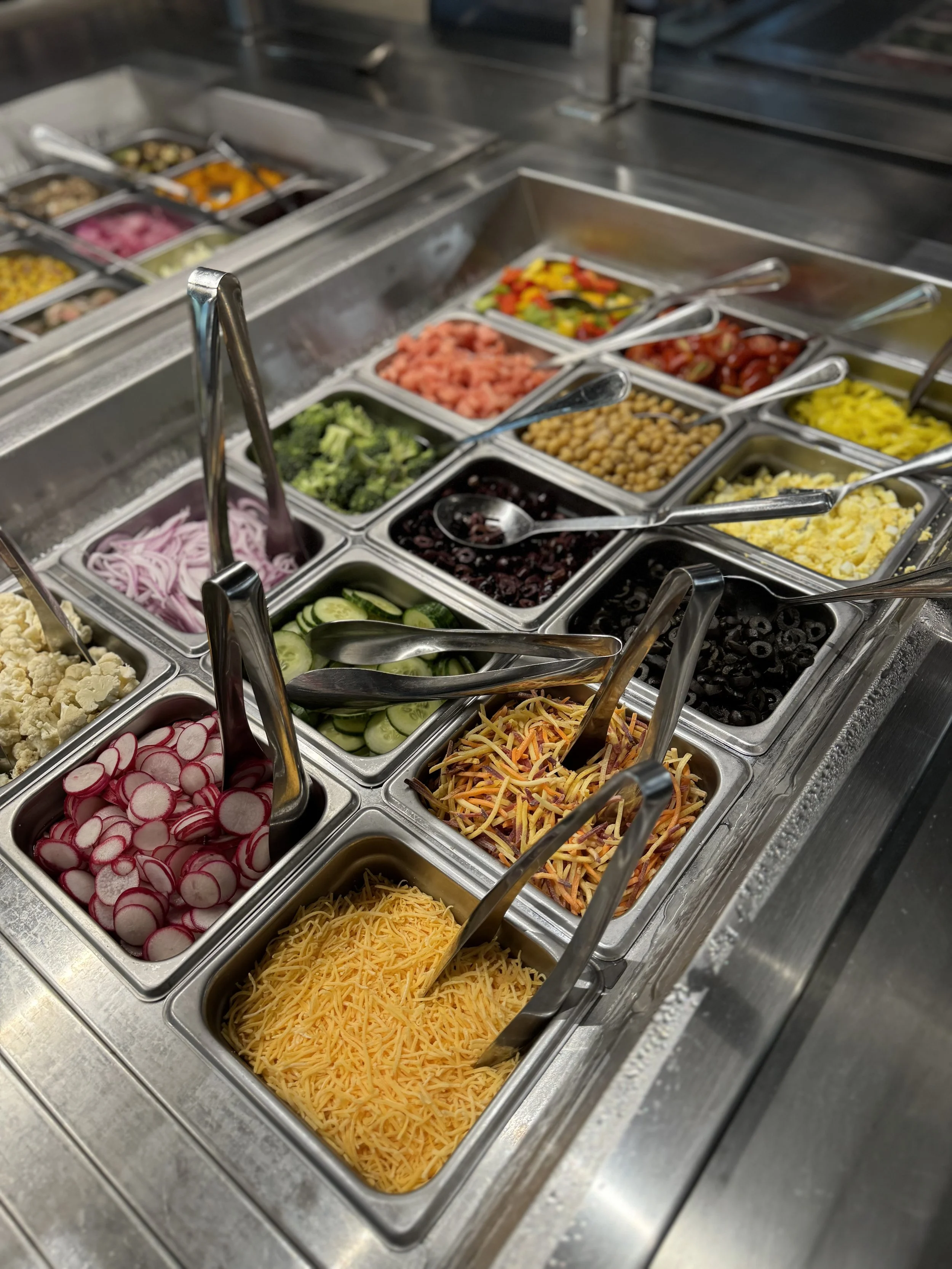 A variety of fresh vegetables and toppings in metal food containers at a salad or taco bar, including sliced radishes, shredded cheese, cucumbers, black olives, shredded carrots, and chopped tomatoes.