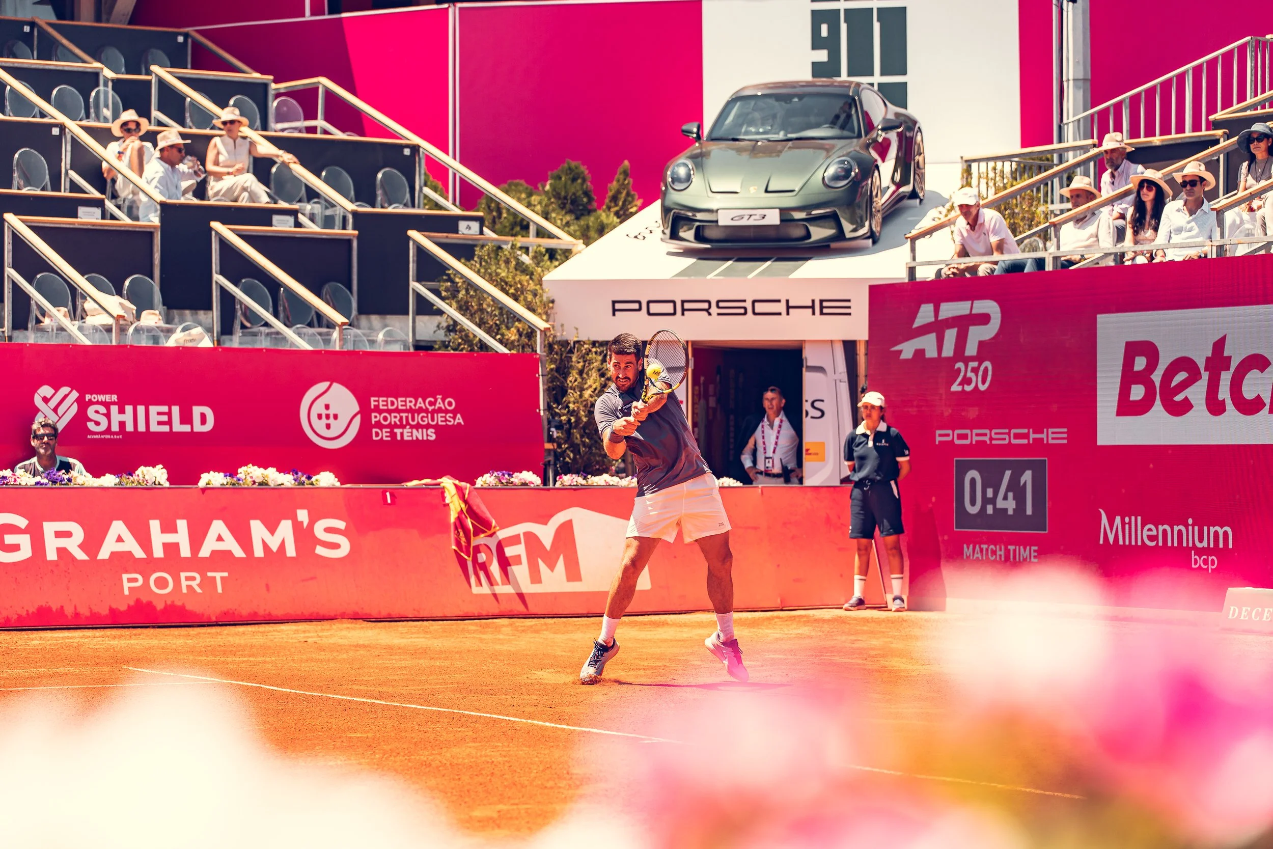 A male tennis player on a clay court during a match, with a tennis ball in mid-air and a tennis racket in his hand, surrounded by bright pink banners and a spectators' stand with people watching.