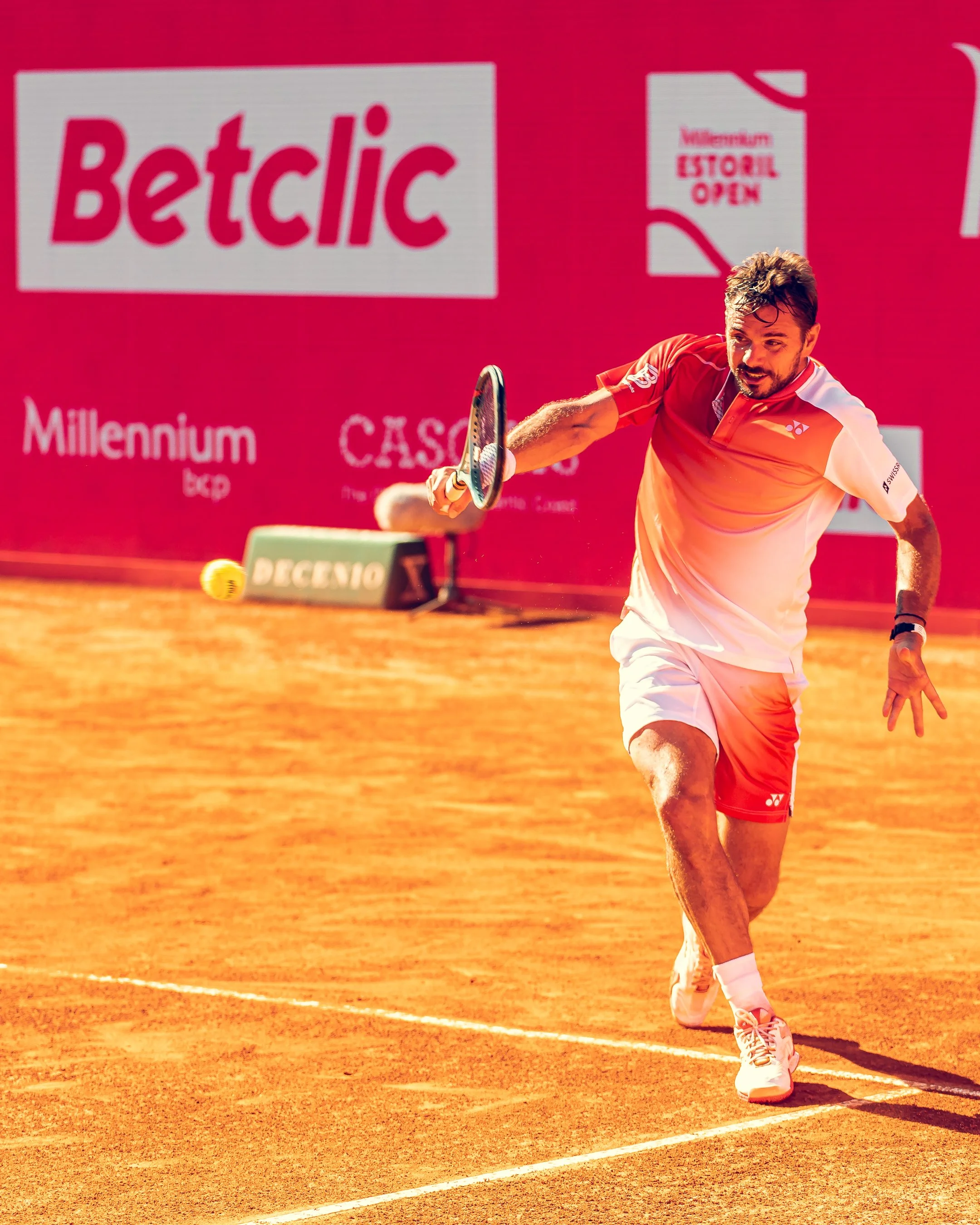 A tennis player in an orange and white outfit swinging a tennis racket on a clay court during a match, with a pink background and sponsor advertisements.