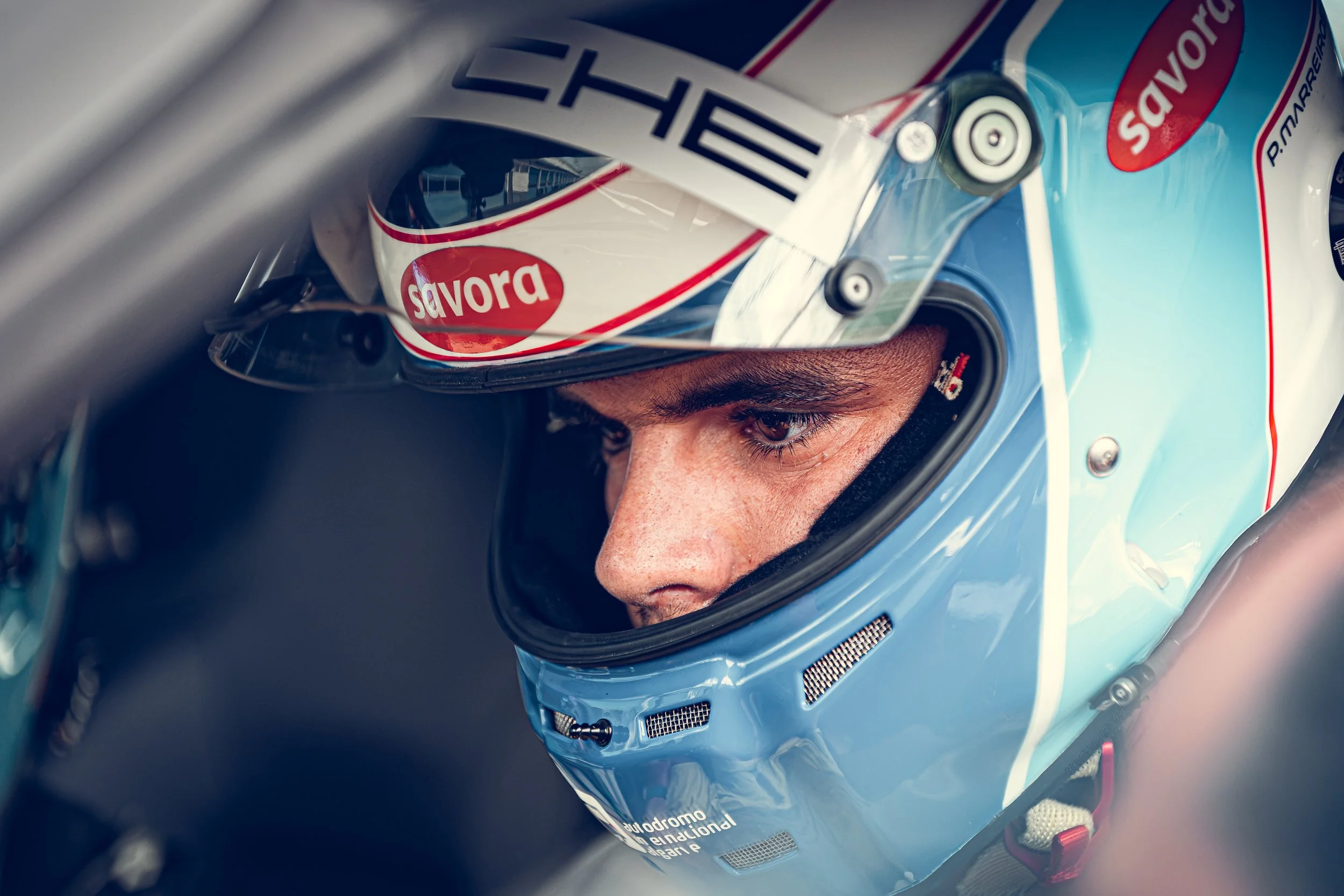 Close-up of a race car driver wearing a blue helmet with a visor, focused intently during a race, with branding and sponsor logos on the helmet and racing suit.
