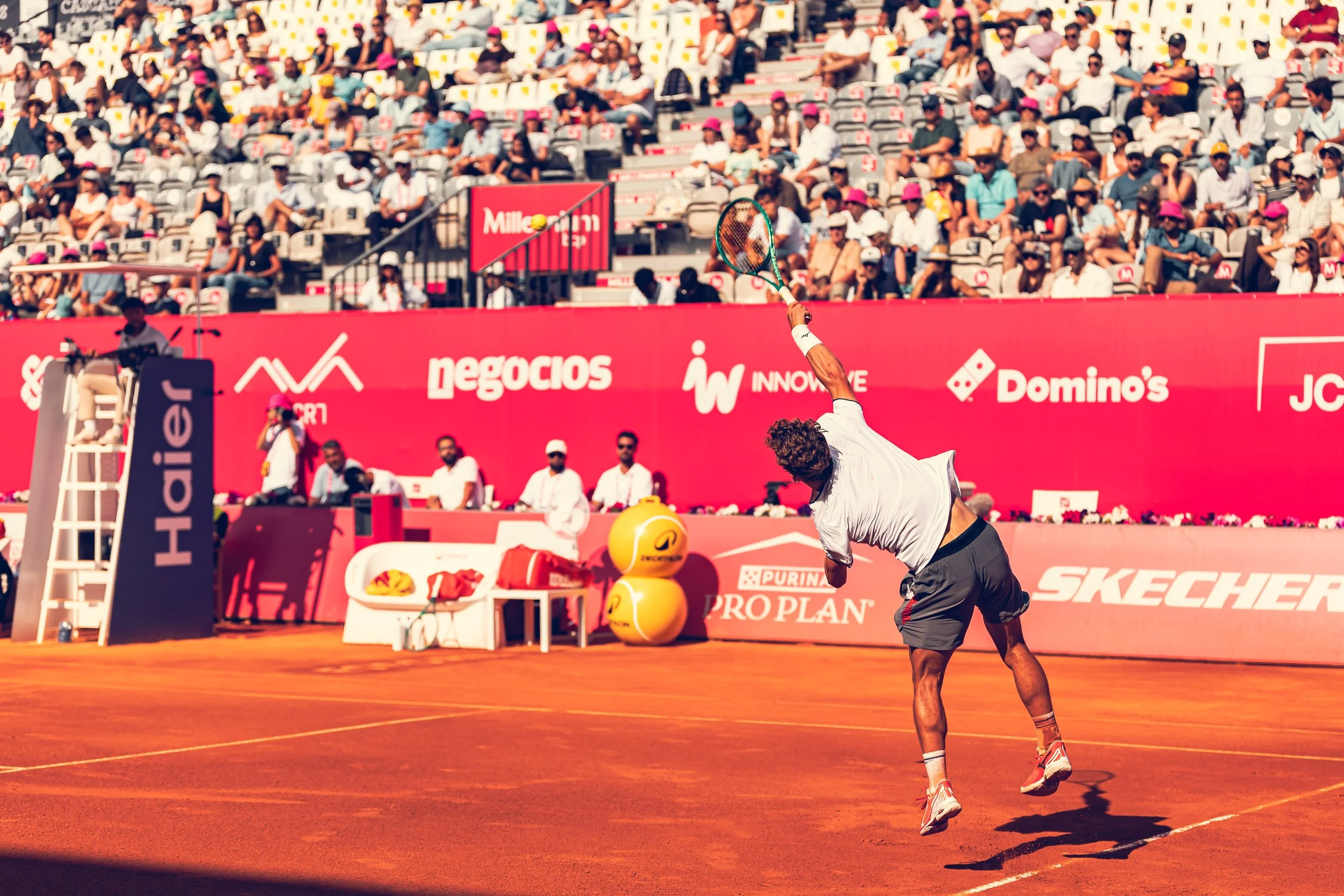 A male tennis player hits a ball with his racket during a match on a clay court, with a large crowd watching from the stands in the background.