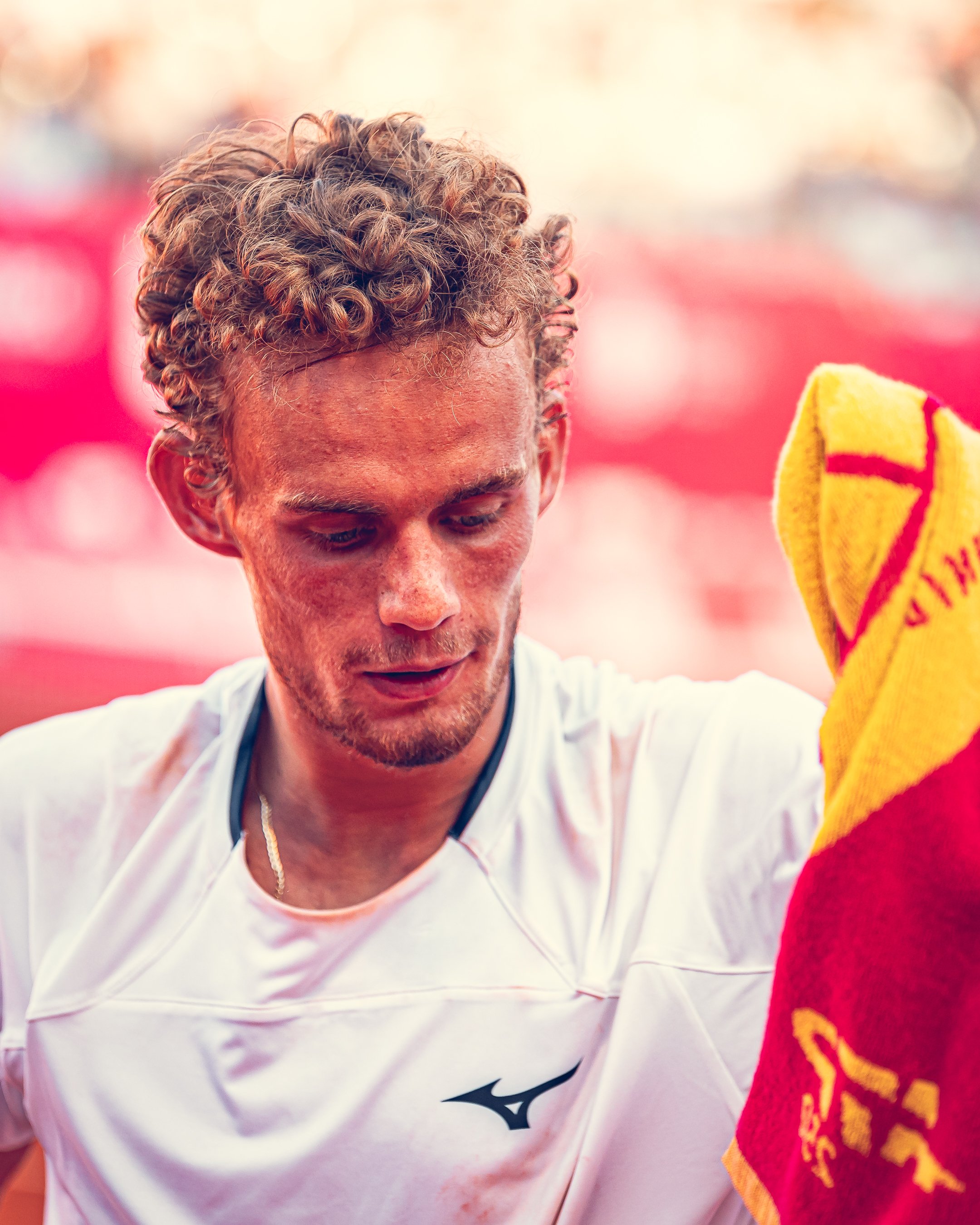 Close-up of a man with curly blond hair wearing a white sports jersey, looking down, with a red and yellow towel held nearby.