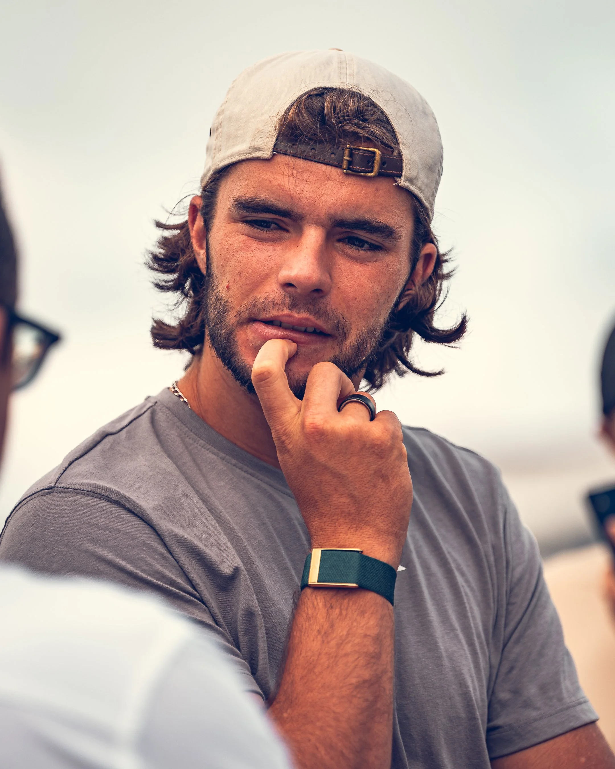 Close-up of a young man with long brown hair, beard, wearing a gray T-shirt, a beige backward cap, and a dark watch, with his hand near his mouth, looking thoughtful.