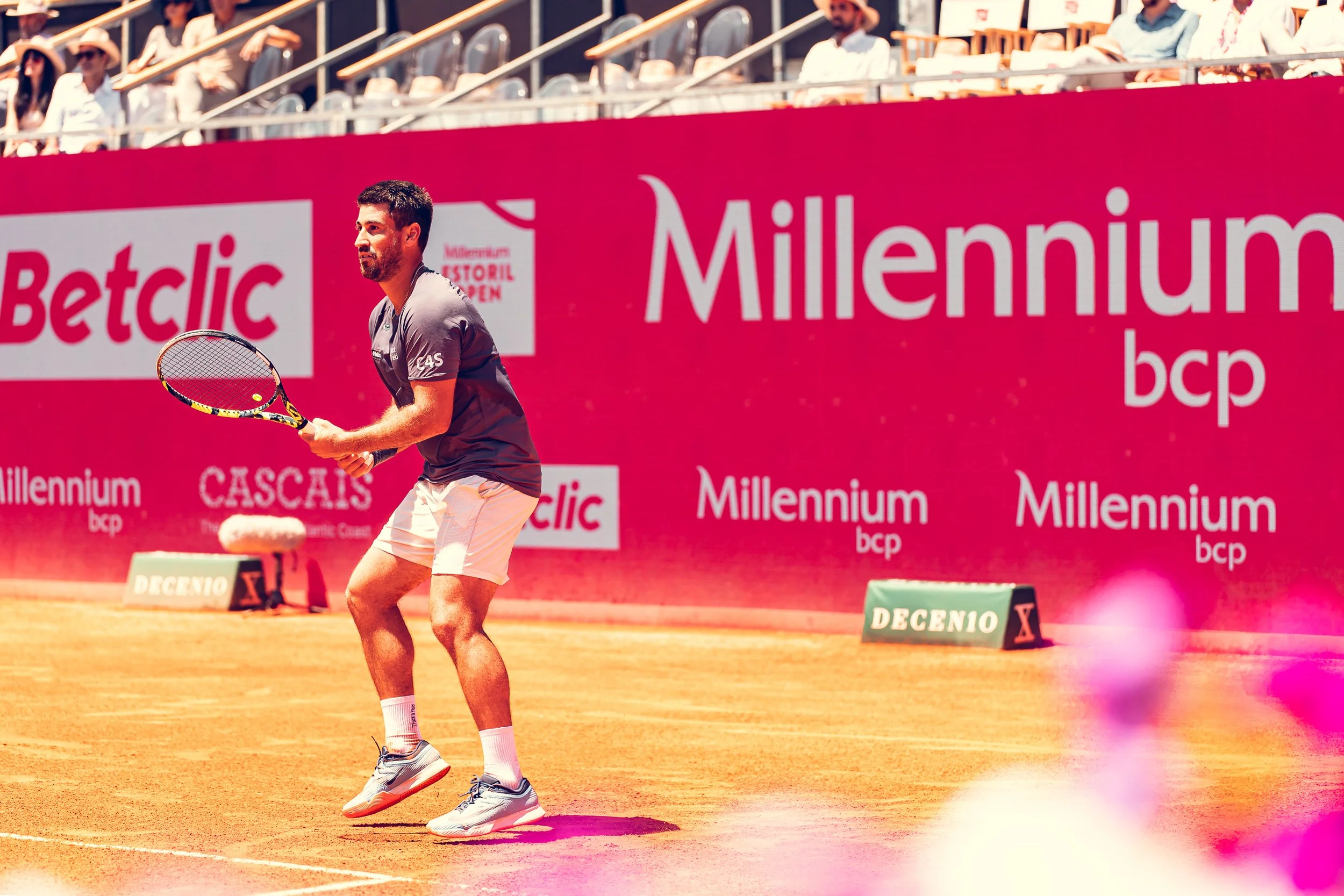 A male tennis player on a clay court during a match, holding a tennis racket, wearing a gray shirt, white shorts, and sneakers, with advertising banners in the background.