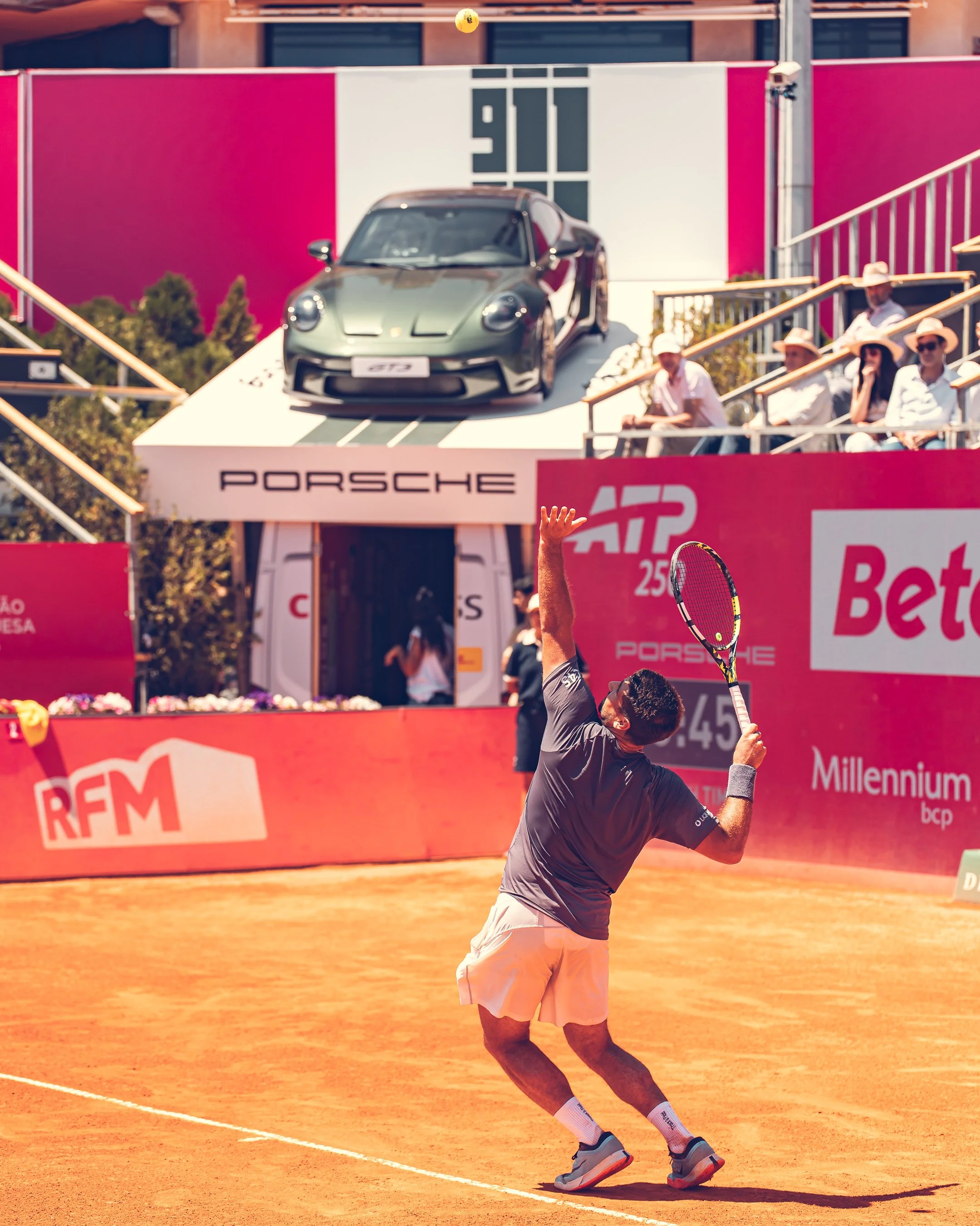 A tennis player in a gray shirt and beige shorts is hitting a tennis ball with a racket on a clay court during a match. Spectators are sitting on bleachers in the background, and banners with sponsor logos are visible.