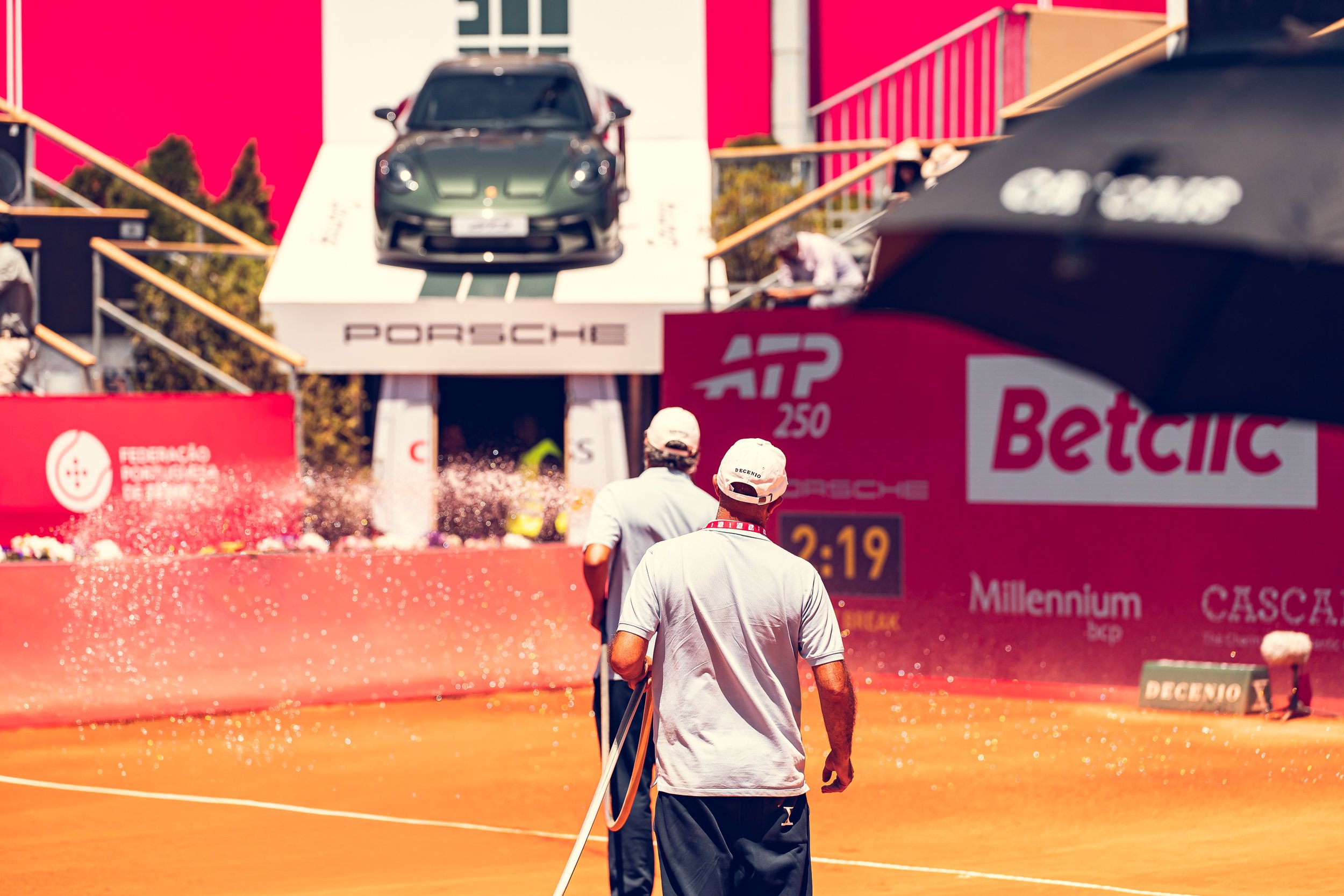 A person walking on a tennis court during a tennis tournament with a Porsche car on an elevated platform in the background.