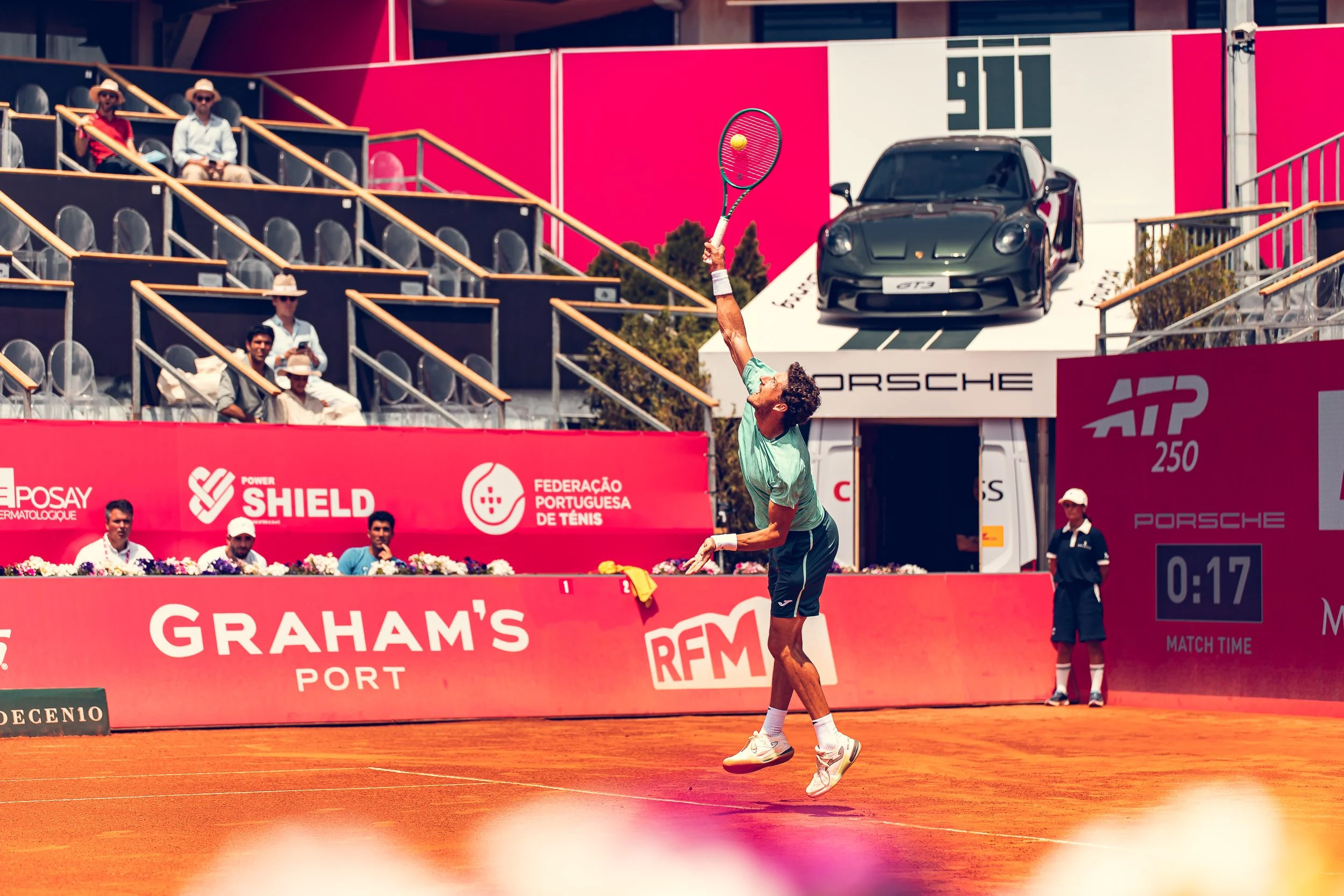 A male tennis player on a clay court, reaching up to hit a tennis ball with his racket during a match, with officials and spectators watching.