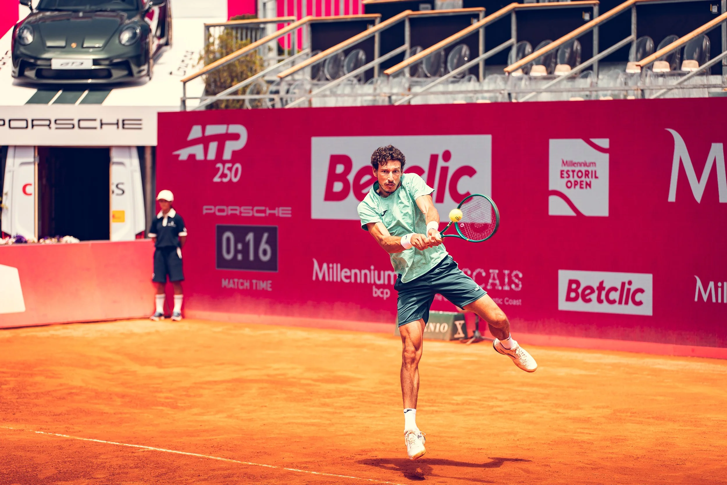 A male tennis player with curly hair and a beard swings his racket to hit a tennis ball on a clay court during a match at the ATP 250 Betclic Millennium Estoril Open.