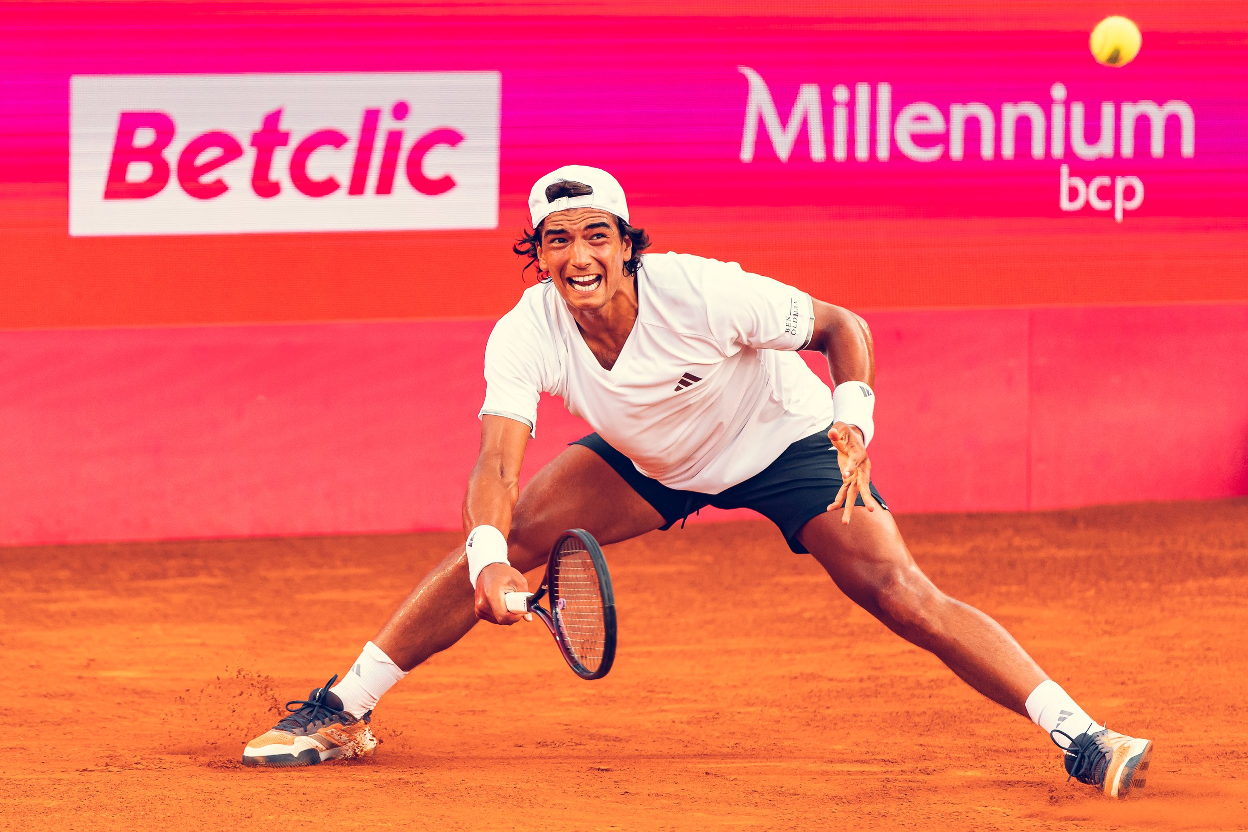 A male tennis player in a white shirt, black shorts, and a white cap playing a match on a red clay court, with a bright pink advertisement background.