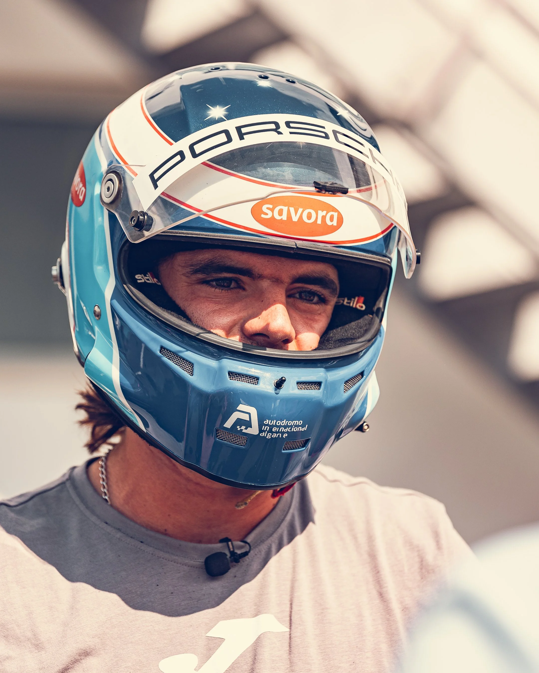 Close-up of a person wearing a racing helmet with 'Porsche' and 'savora' logos, visible eyes, and part of a light gray t-shirt.