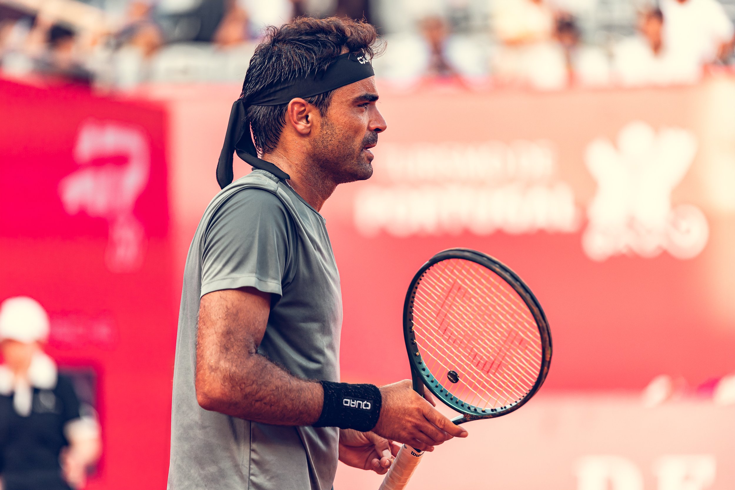 A male tennis player standing on a tennis court, holding a tennis racket, wearing a black headband and a grey sports shirt, with a blurred tennis court background.
