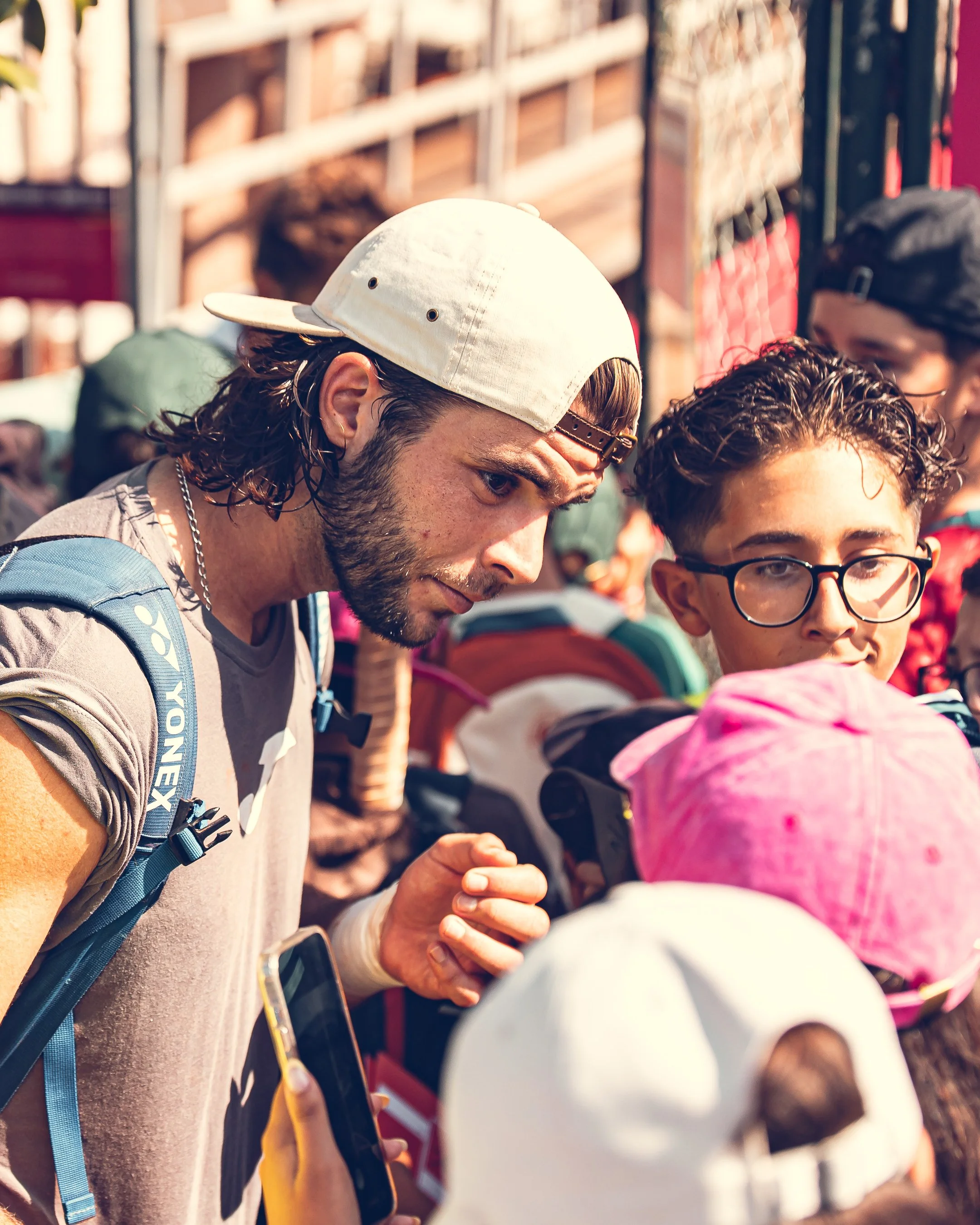 People gathered outdoors, some wearing hats, with a man in the foreground wearing a white cap and a backpack, and a woman next to him wearing glasses and a pink or purple head covering.