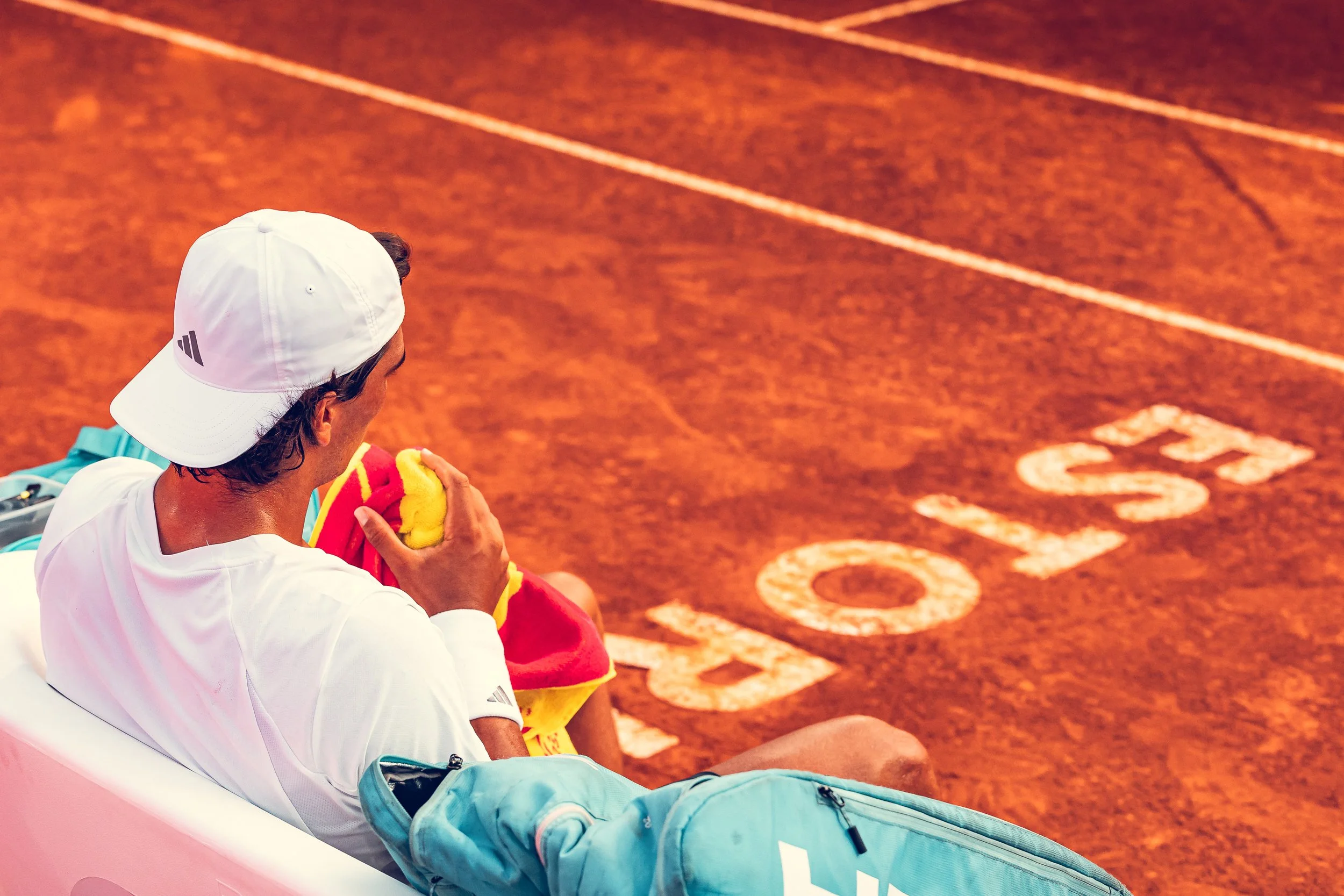 A tennis player sitting on the bench of a clay tennis court, holding a towel near his face, with a white cap and a blue backpack.