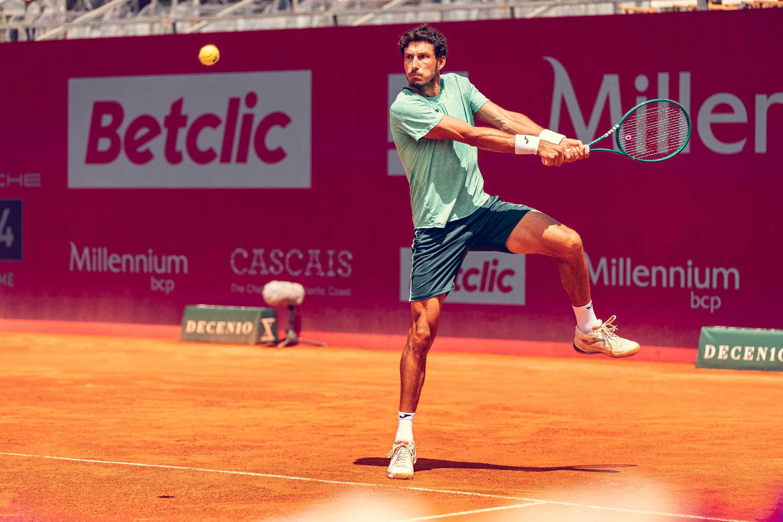 A male tennis player in a green shirt and black shorts preparing to hit a tennis ball on a clay court. The background features advertising banners with the words 'Betclic' and 'Millennium'.