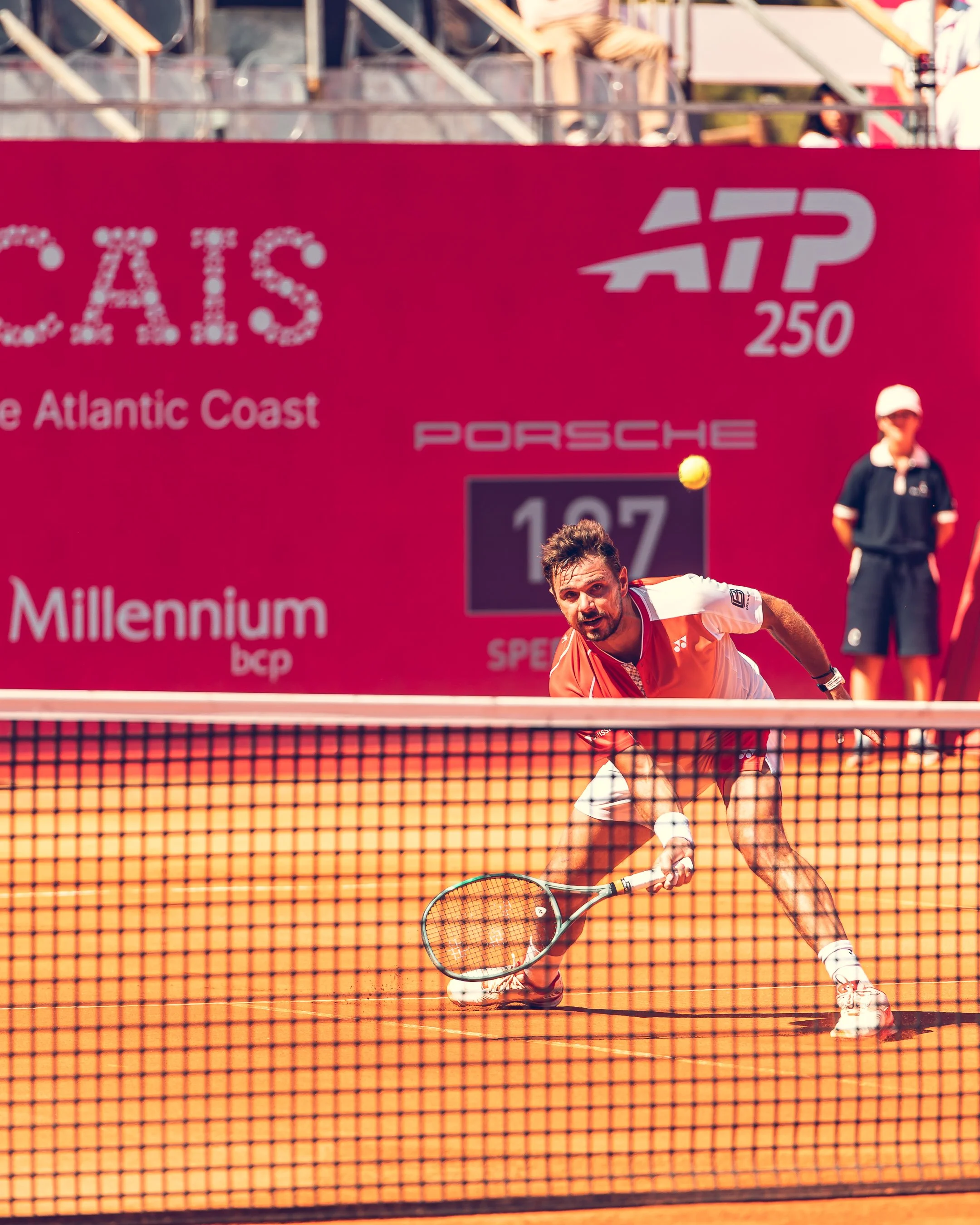 A male tennis player wearing a white and orange outfit, playing on an orange clay court during a match. He is holding a tennis racket and appears to be reaching to hit the ball. There is a pink banner in the background with advertisements and a crowd