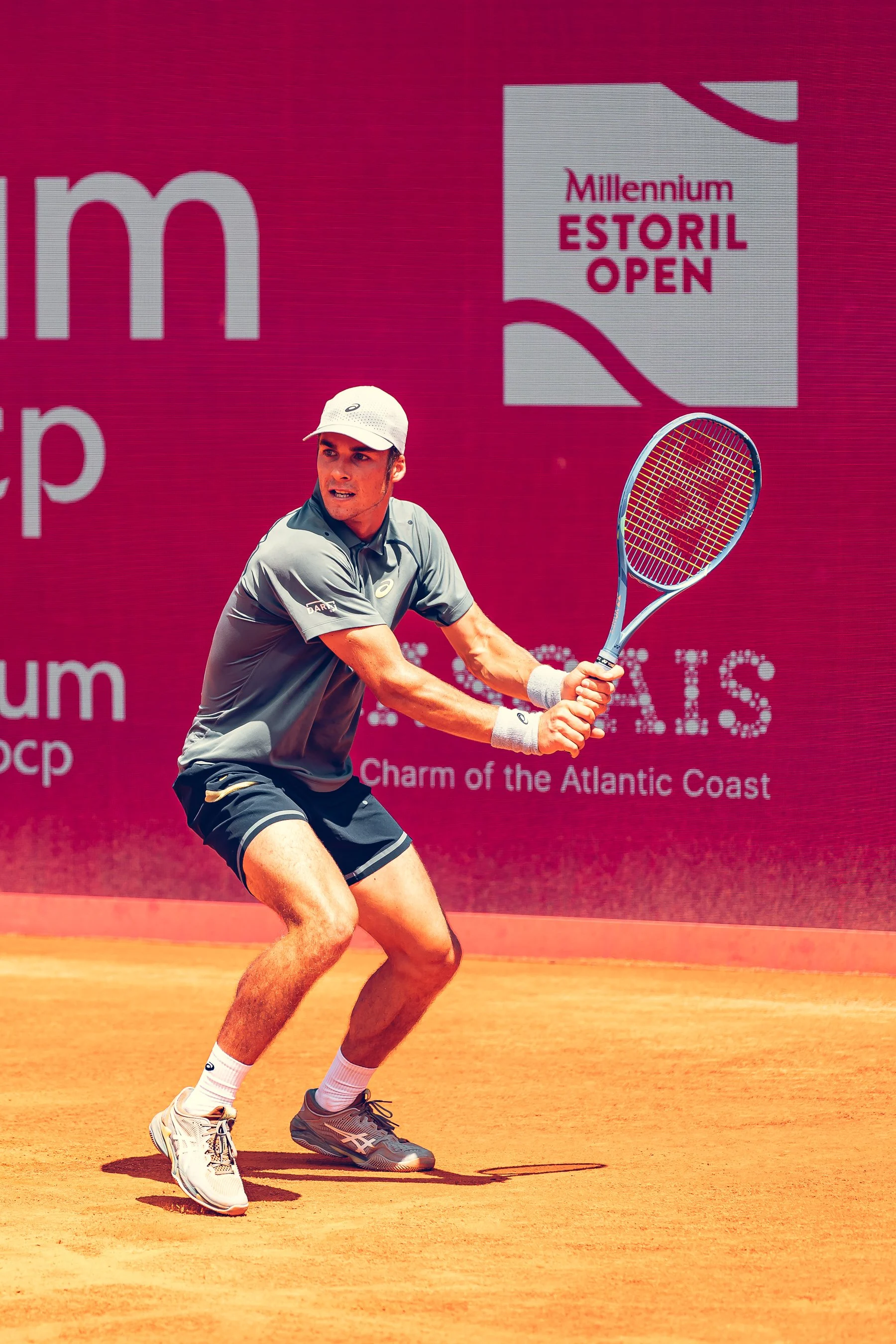 A male tennis player in gray athletic clothing, white hat, and tennis shoes, playing on a clay court at the Millennium Estoril Open with a pink tournament banner in the background.