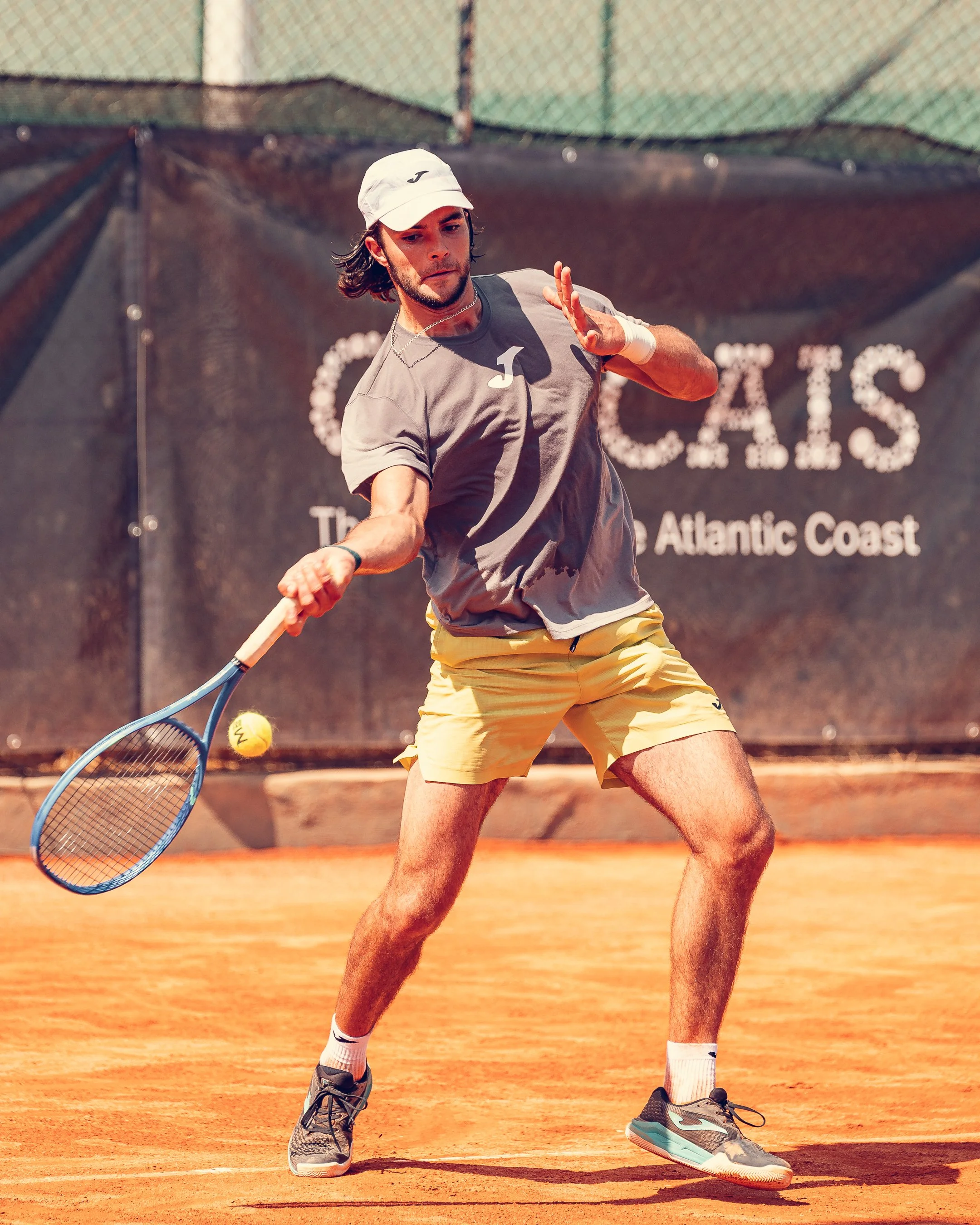 A young man playing tennis on a clay court, dressed in a white cap, gray t-shirt, yellow shorts, and gray athletic shoes, preparing to hit a tennis ball with his racket.