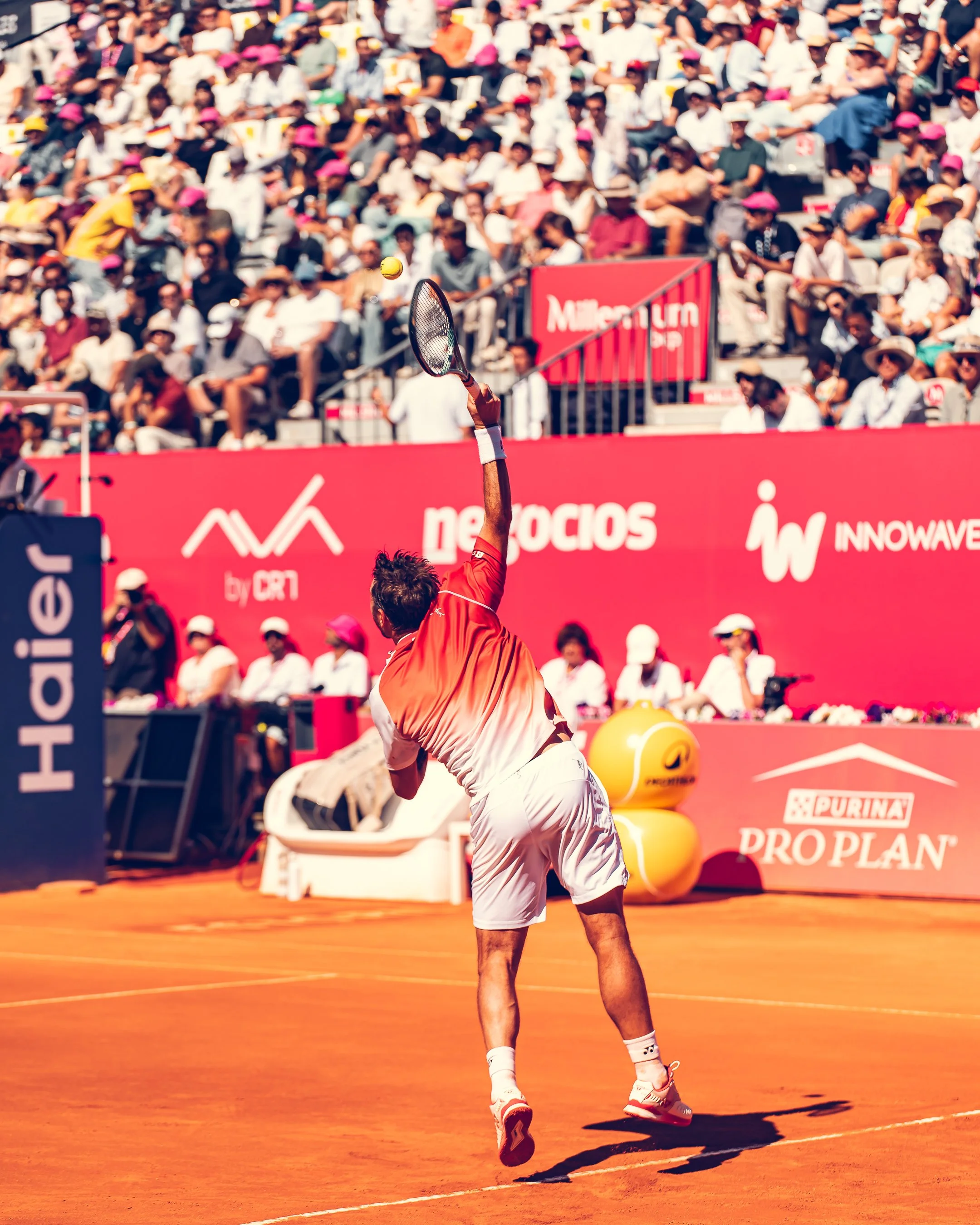 A tennis player in red and white attire hitting a tennis ball on a clay court, with a large audience in the background watching the match.