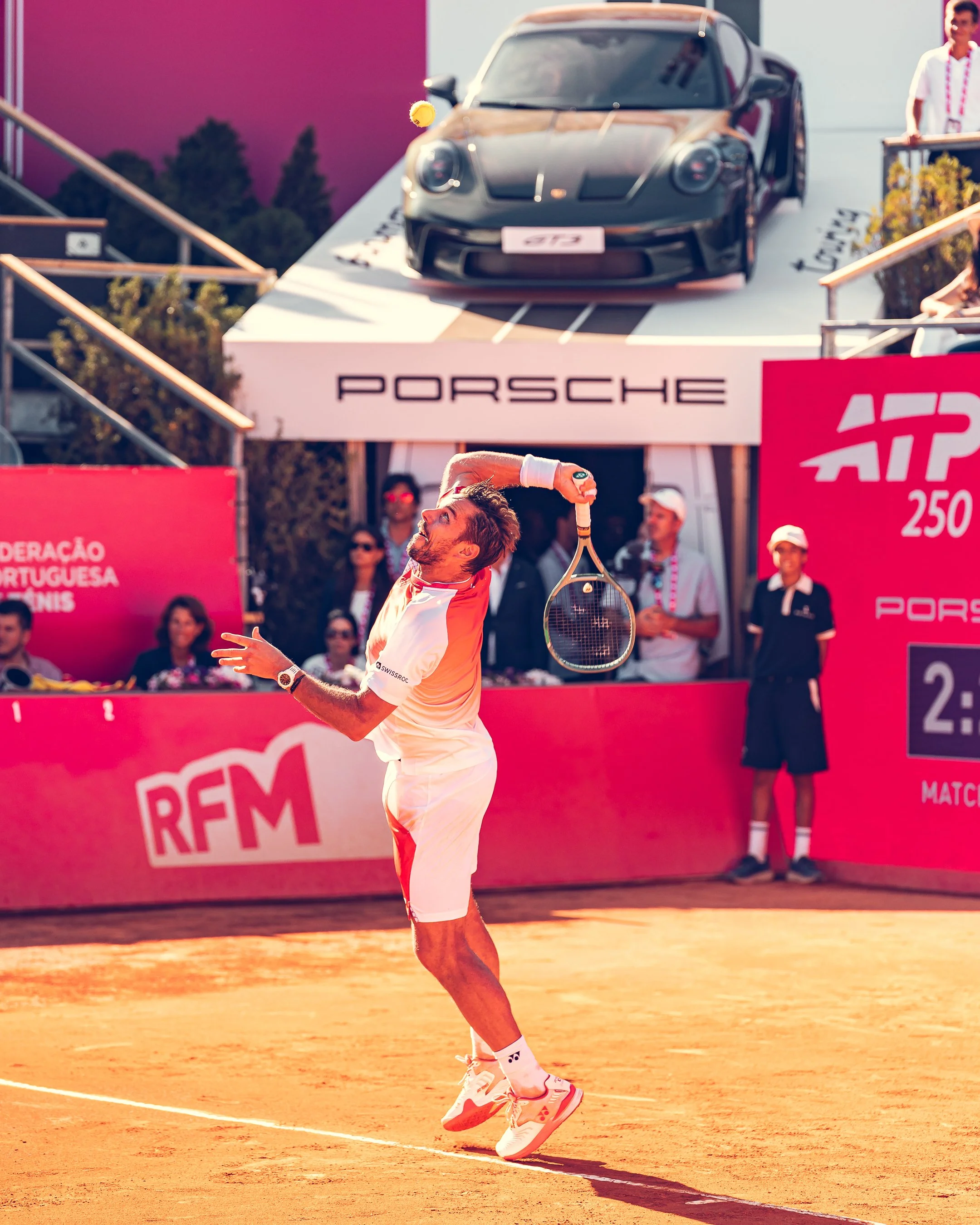 A tennis player is on a clay court, preparing to serve, wearing a white and orange uniform and Yonex tennis shoes. A tennis ball is in midair above his racket. In the background, there is a Porsche car on a platform and several spectators watching.