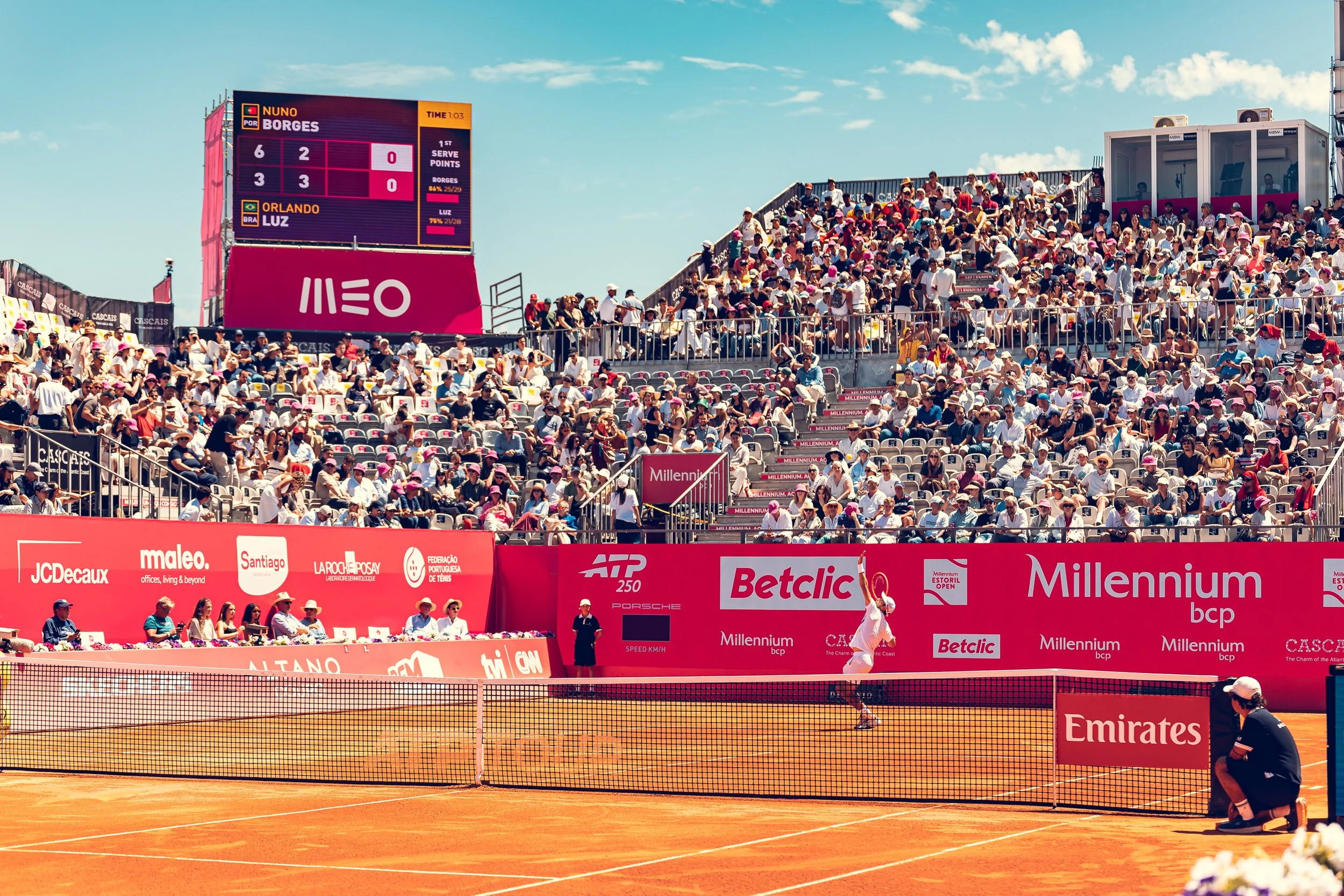 Spectators seated in a tennis stadium, with a tennis match underway on the court. A digital scoreboard shows the players' names, Nuno Borges and Orlando Luz, with scores 6-3 and 2-3 respectively. There are various sponsor banners along the perimeter 