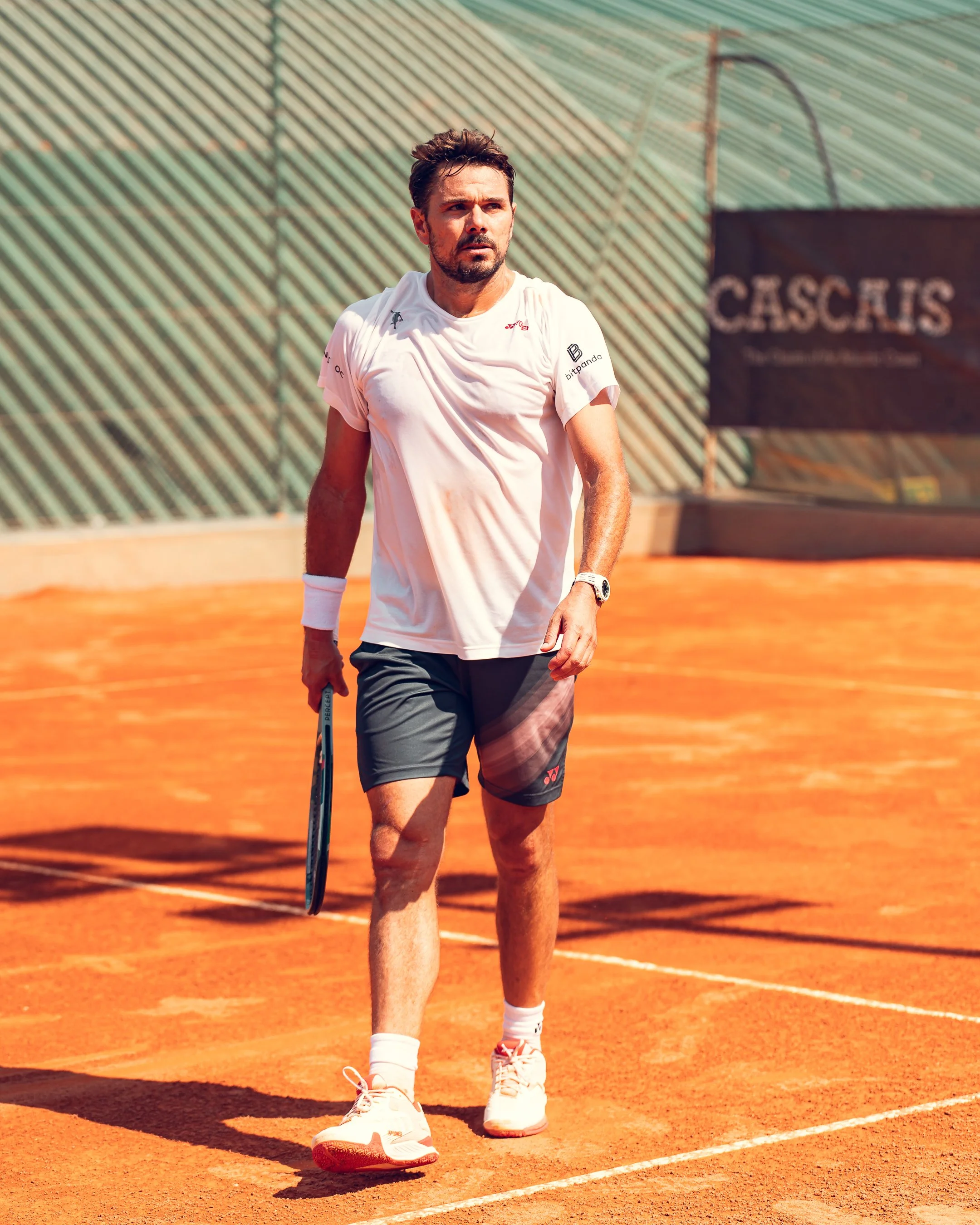 A male tennis player on a clay court, dressed in a white t-shirt, dark shorts, and white shoes, holding a tennis racket in his right hand.