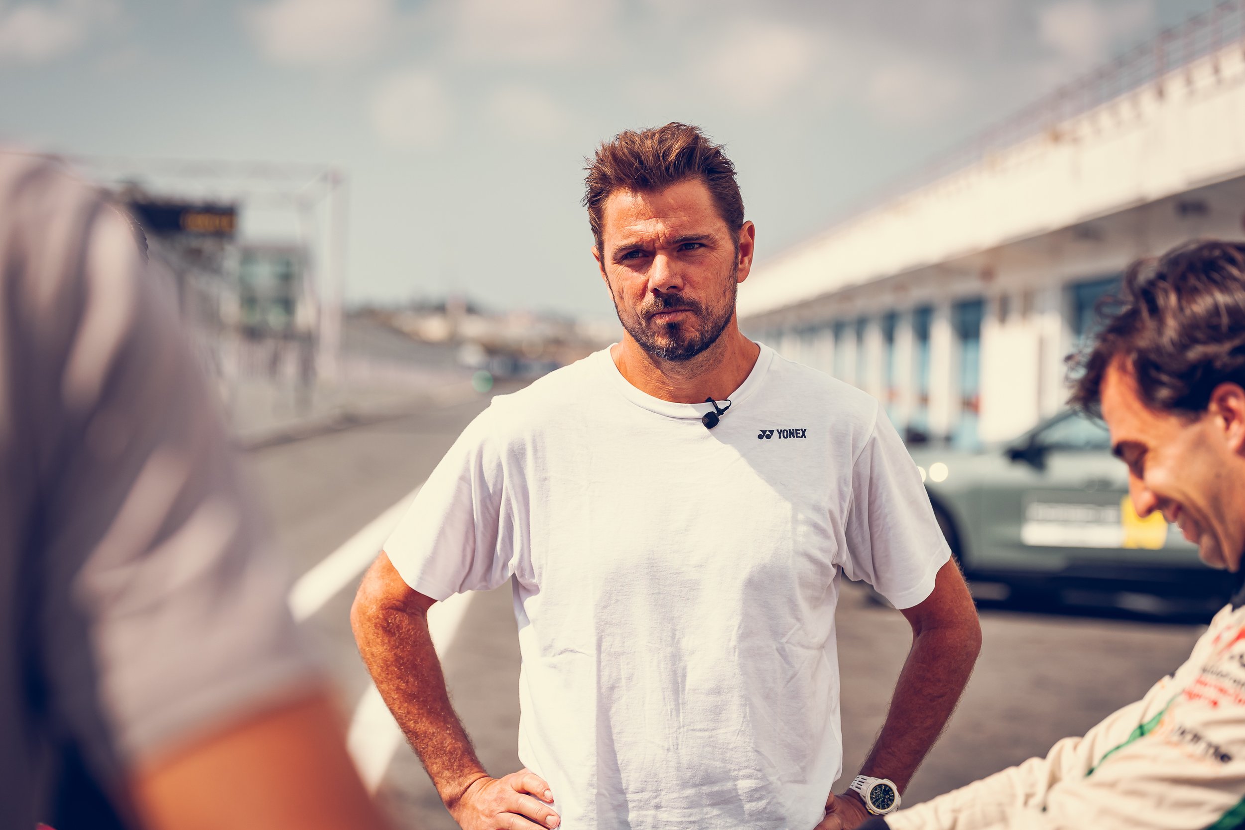 A man with brown hair and a beard, wearing a white T-shirt and a watch, stands outdoors with his hands on his hips. He appears to be in conversation with people whose partial faces are visible in the foreground. The background shows a bridge and a pa