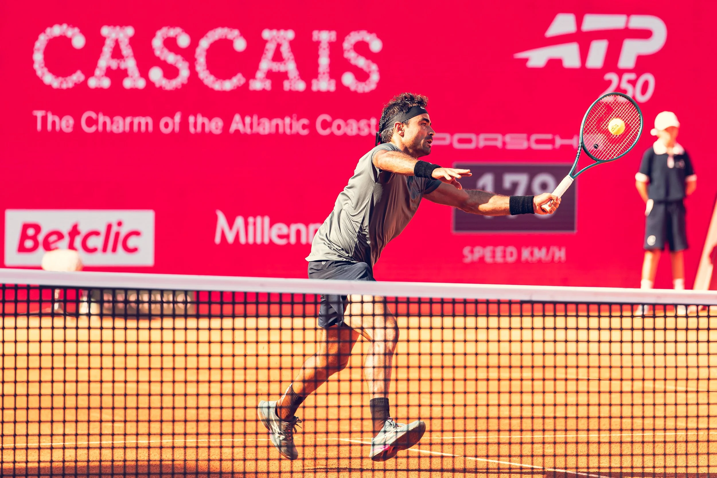 A male tennis player in a gray shirt and black shorts is playing tennis on a clay court, hitting the ball with a racket. A net is visible in the foreground. The background has pink signage with the words "CASCA" and various logos.