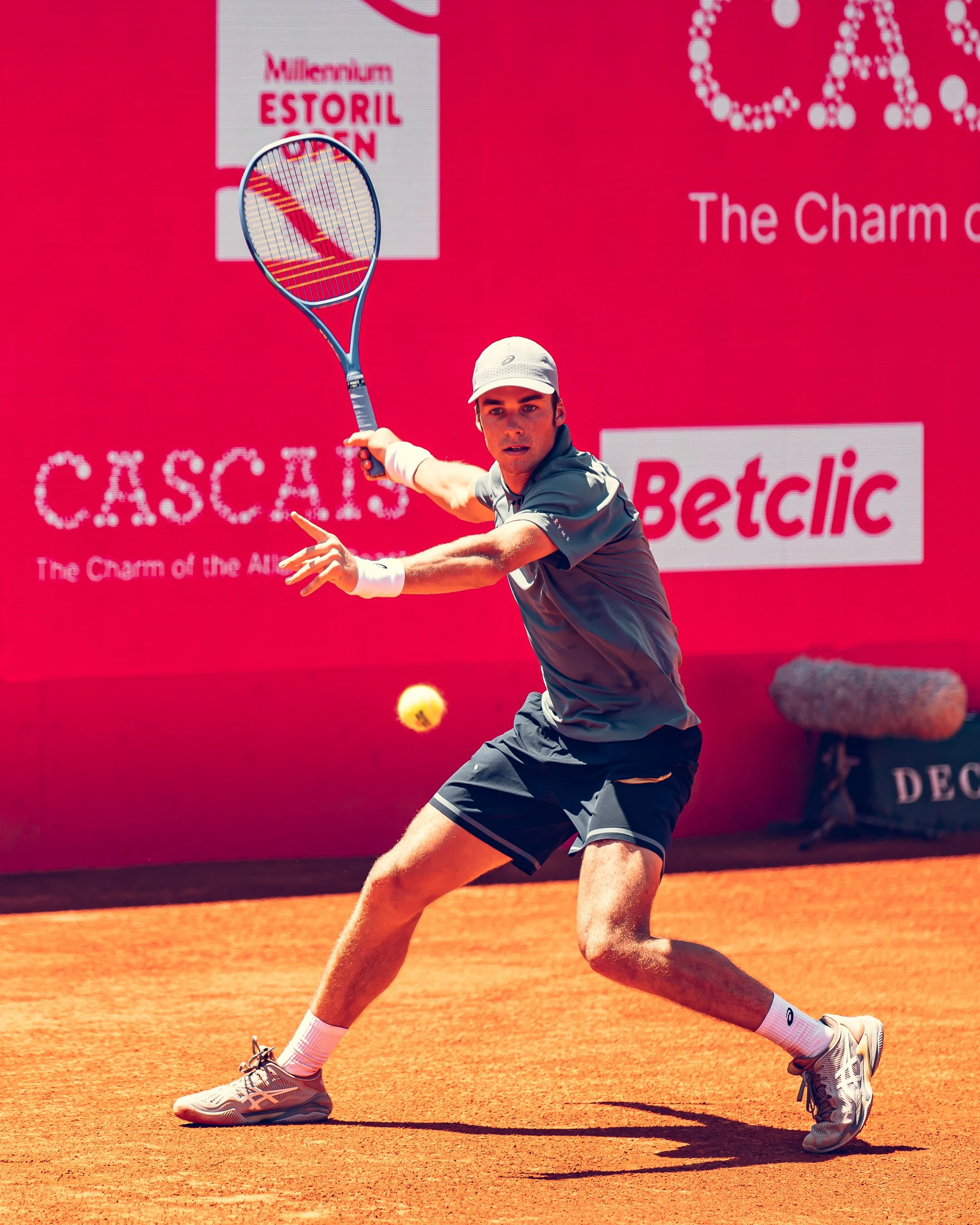 A male tennis player in a gray shirt, black shorts, and a light-colored cap preparing to hit a tennis ball on a clay court.