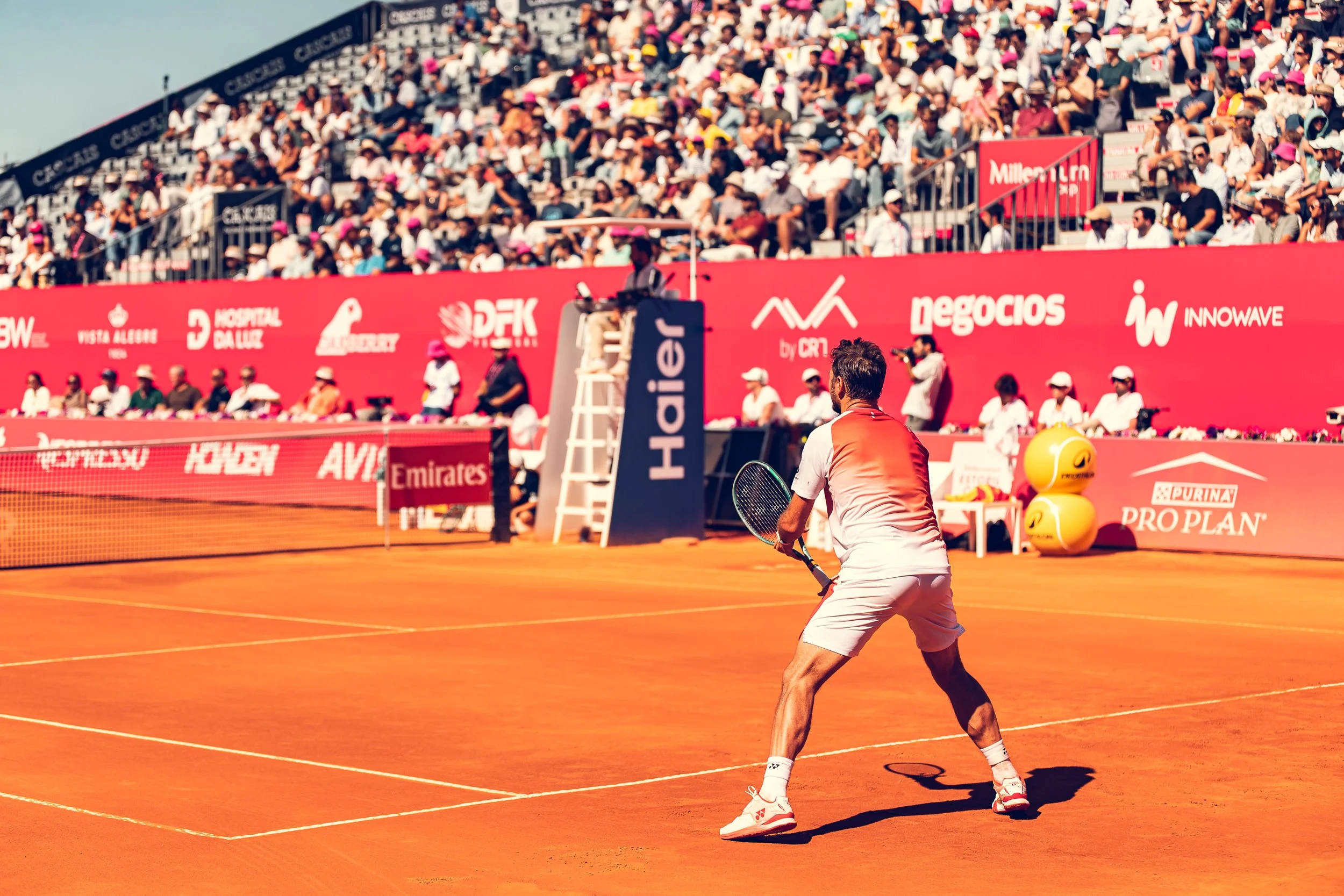 A male tennis player in white attire preparing to hit a ball on an orange clay tennis court during a professional match. Spectators watch from the stands in the background, and there are advertising banners along the perimeter of the court.