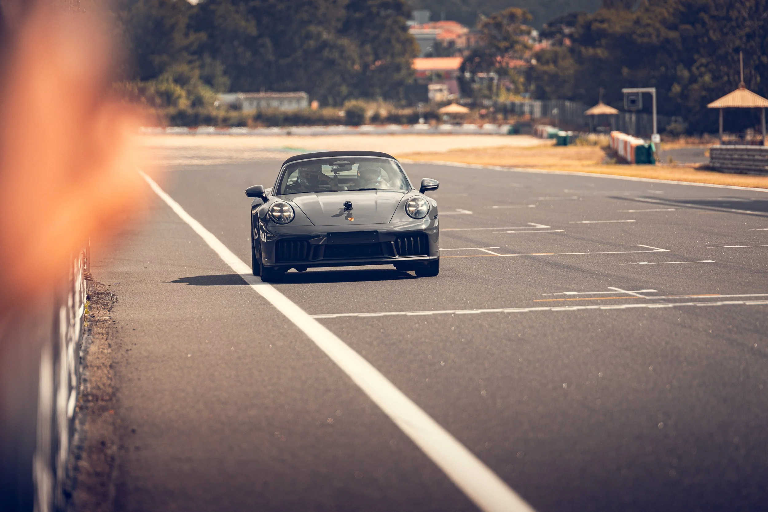 A gray sports car driving on a racetrack, with a blurred foreground of a barrier and trees in the background.
