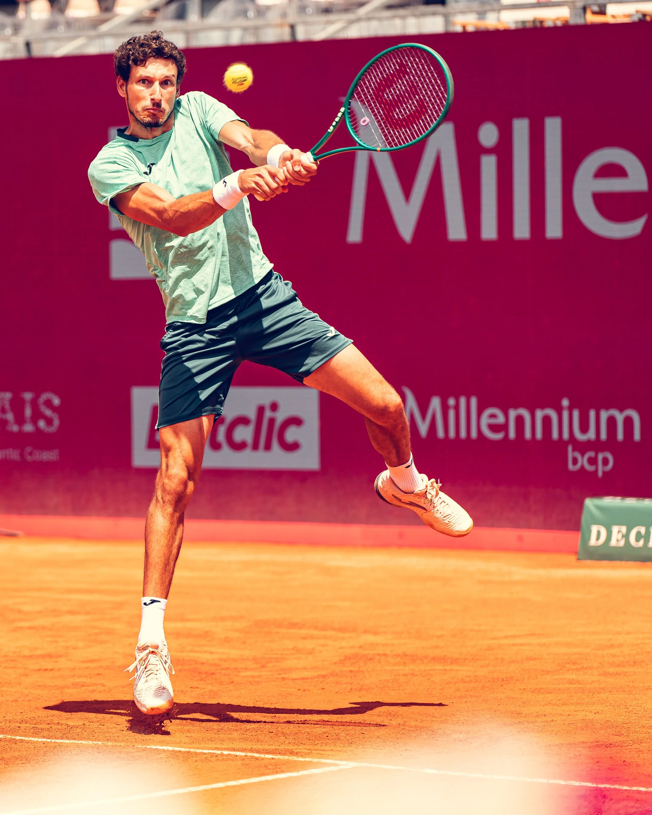 A tennis player is hitting a tennis ball with a racket on a clay court. He is wearing a light green shirt, dark shorts, white wristbands, and white sneakers. The background features advertisements and seating stands.