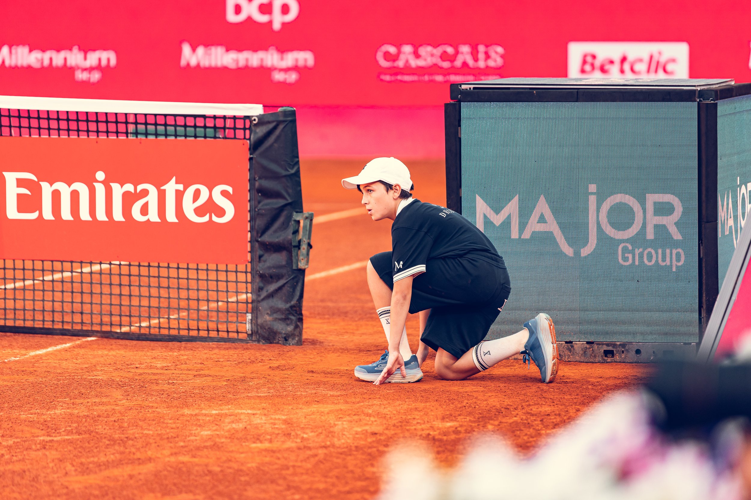 A young male tennis player kneeling on a clay court, preparing to serve, with tennis net and advertising banners in the background.