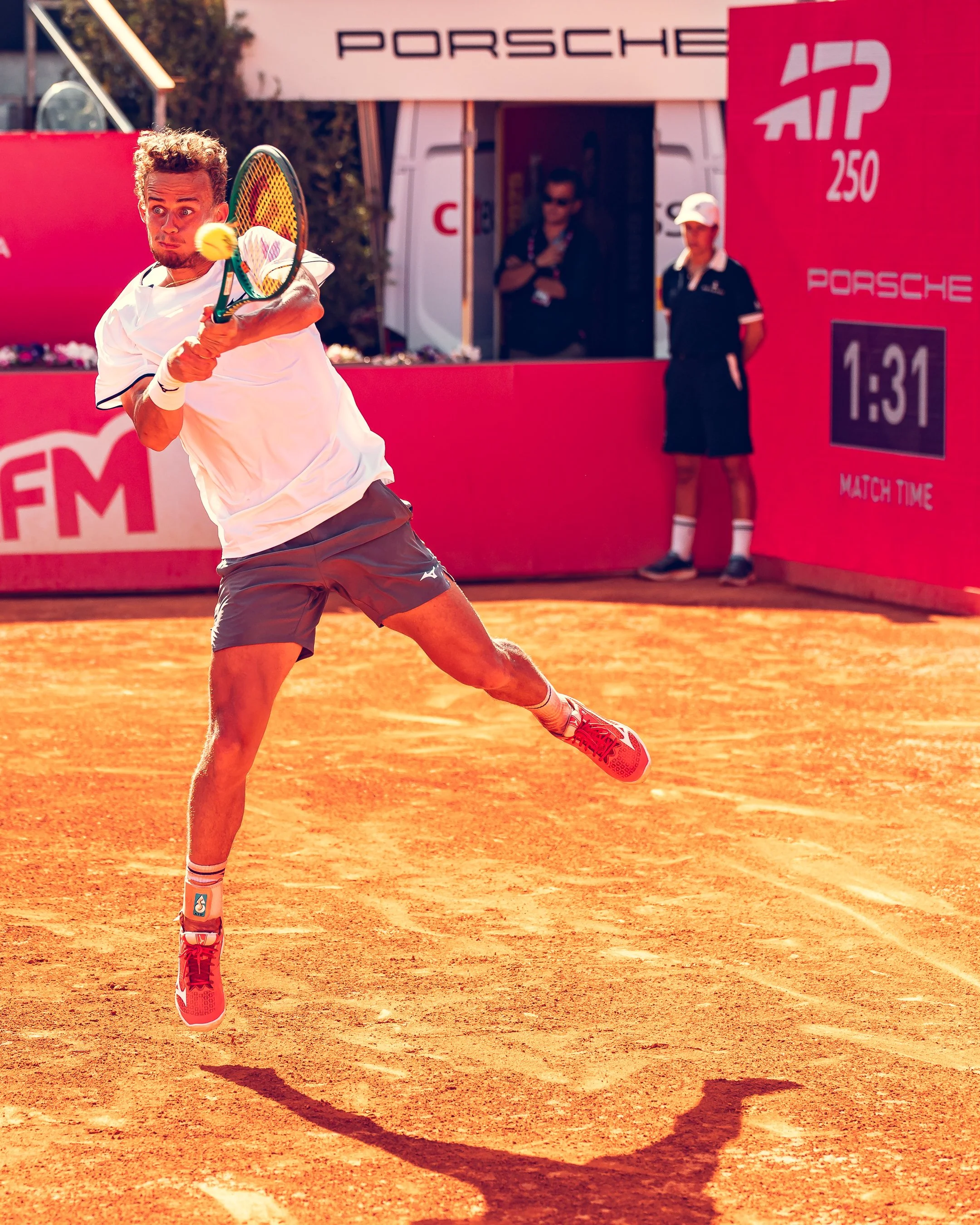 A male tennis player in mid-swing hitting a tennis ball on a clay court, with a red and white banner showing sponsors and time in the background.