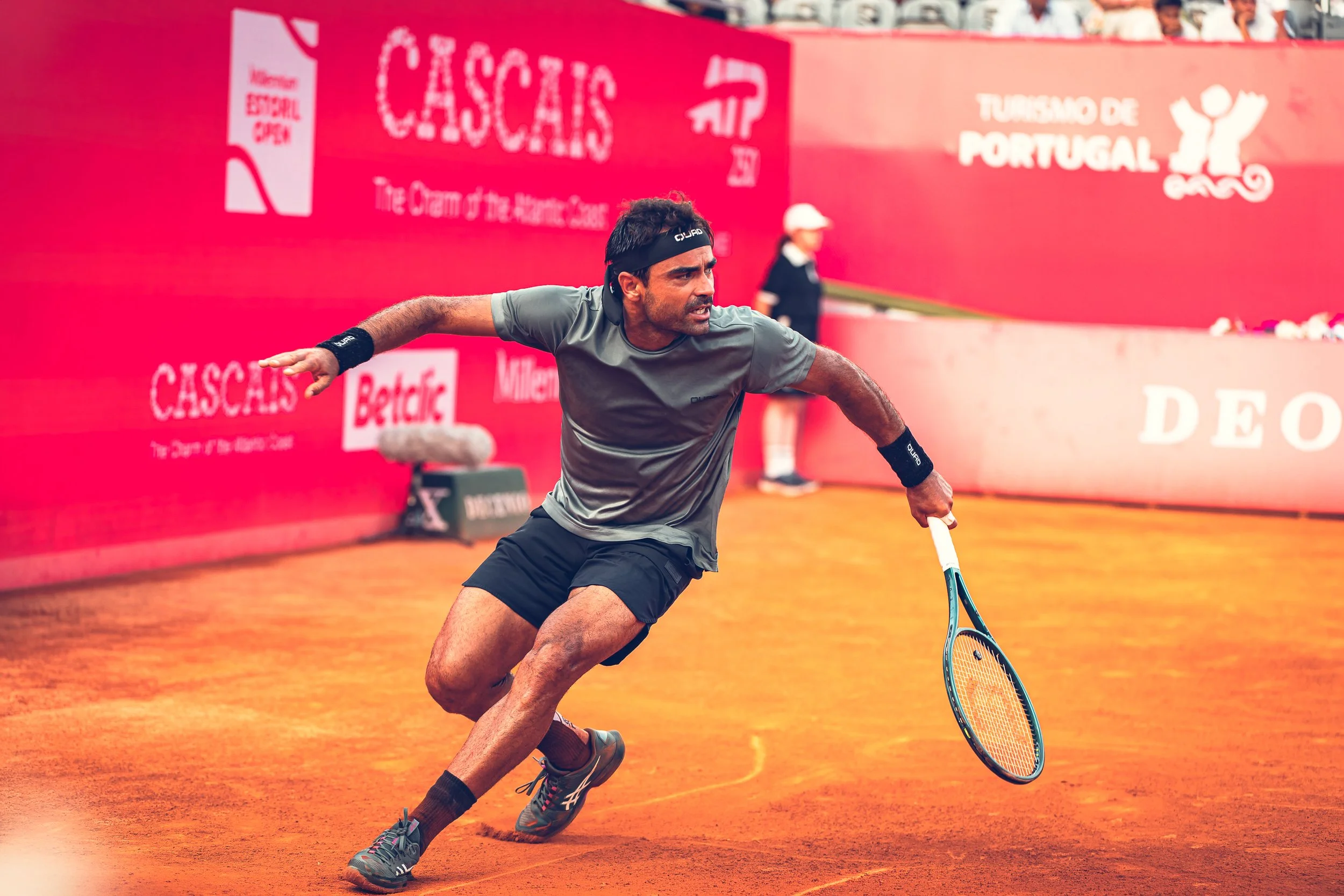 A male tennis player in motion on a clay court, wearing a gray shirt, black shorts, and a black headband, with a blue tennis racket, during a match at the Cascais tournament in Portugal.