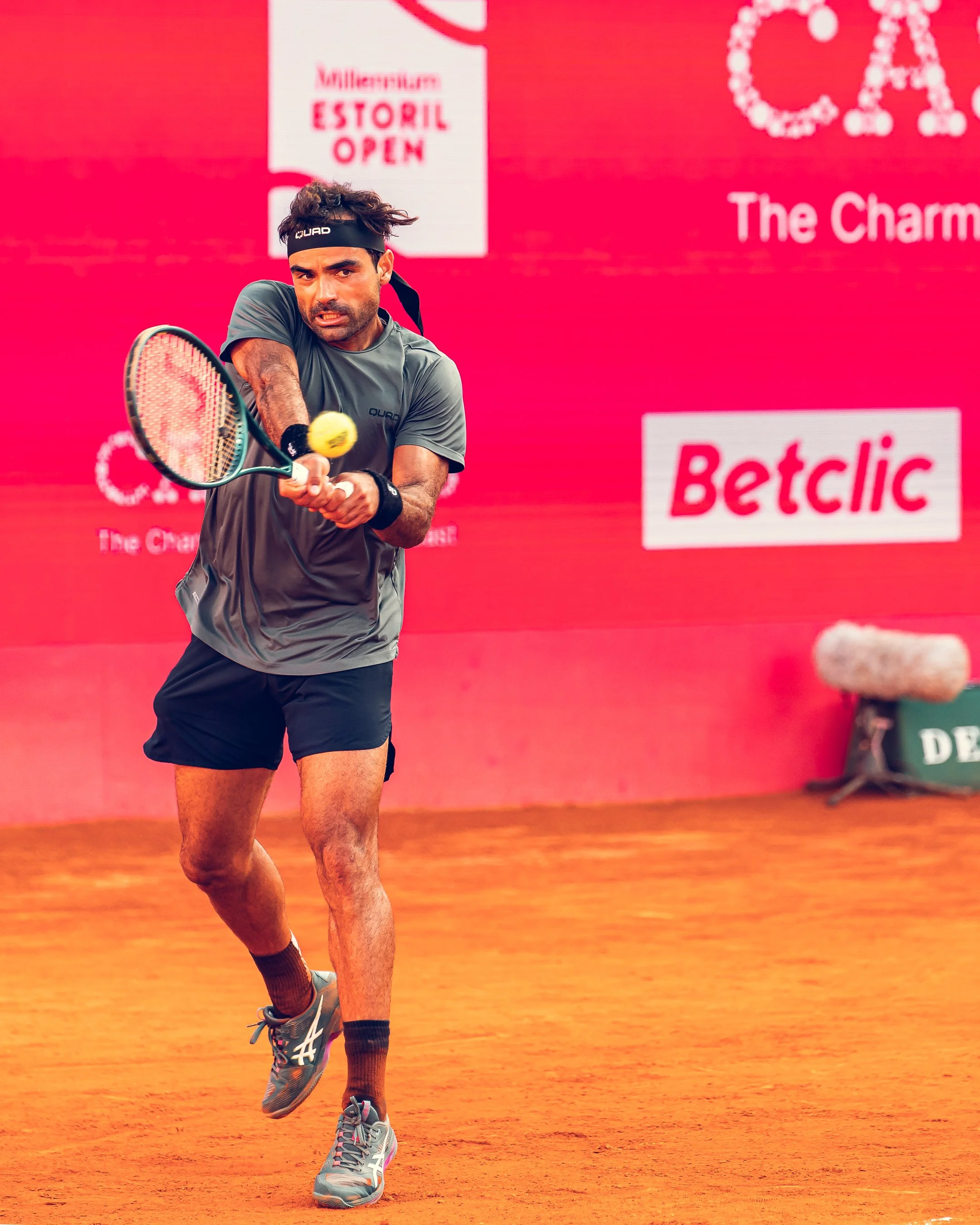 A male tennis player hitting a ball on a clay court during a match, with a bright pink backdrop featuring sponsor logos and event signs.