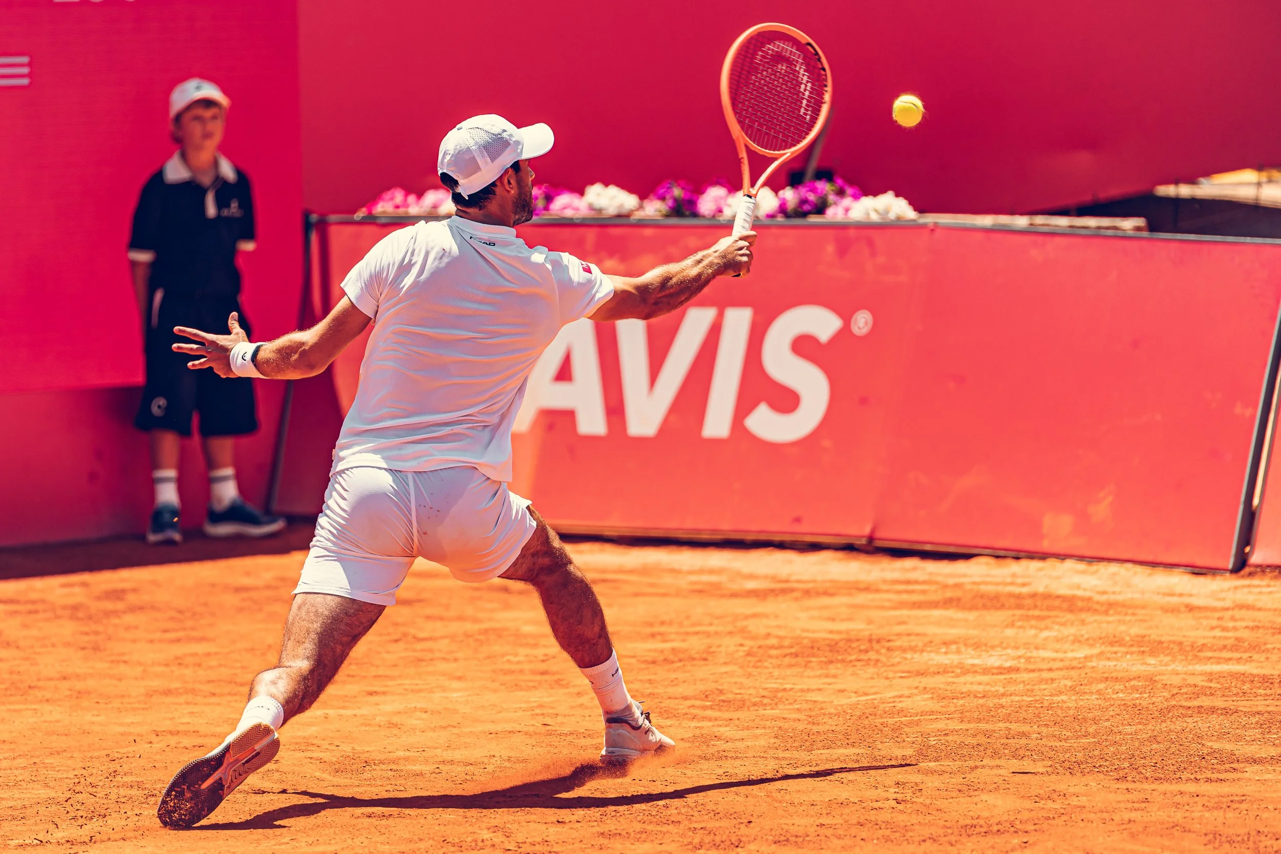 A tennis player in white attire and a white cap hitting a tennis ball with a racket on a clay court, with a ball girl or official standing in the background, and a red backdrop with the word 'AVIS'.