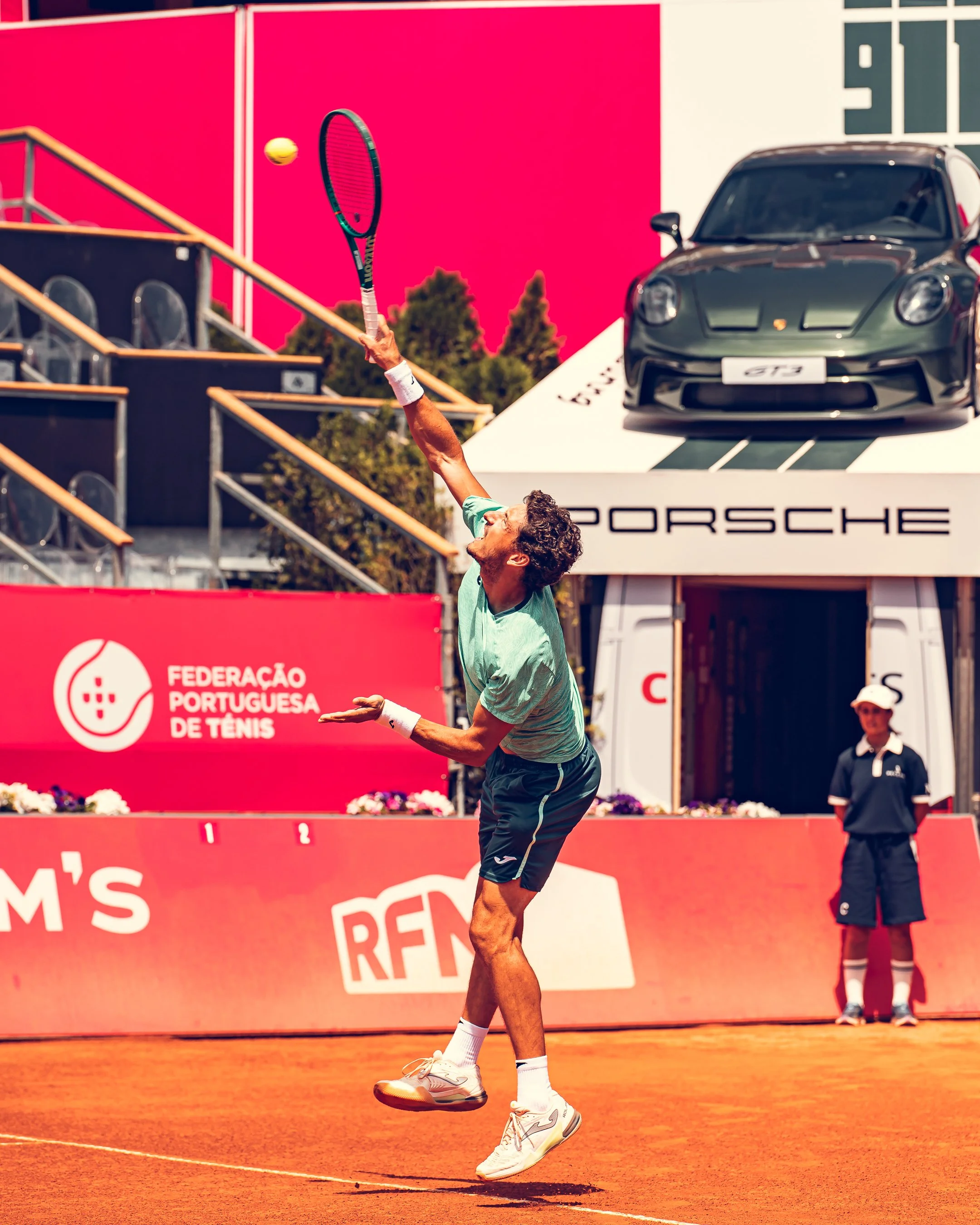 A tennis player serves on a clay court during a match, with a digital scoreboard, a Porsche car display, and a person in a navy outfit in the background.