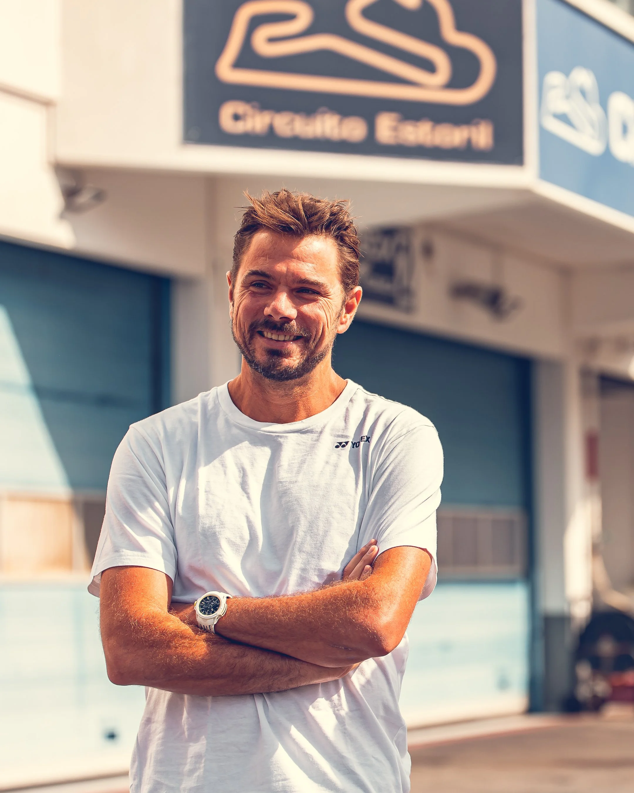 A smiling man with short brown hair, a beard, and a mustache, wearing a white T-shirt and a watch, standing outdoors near a body of water with a building and signs in the background.