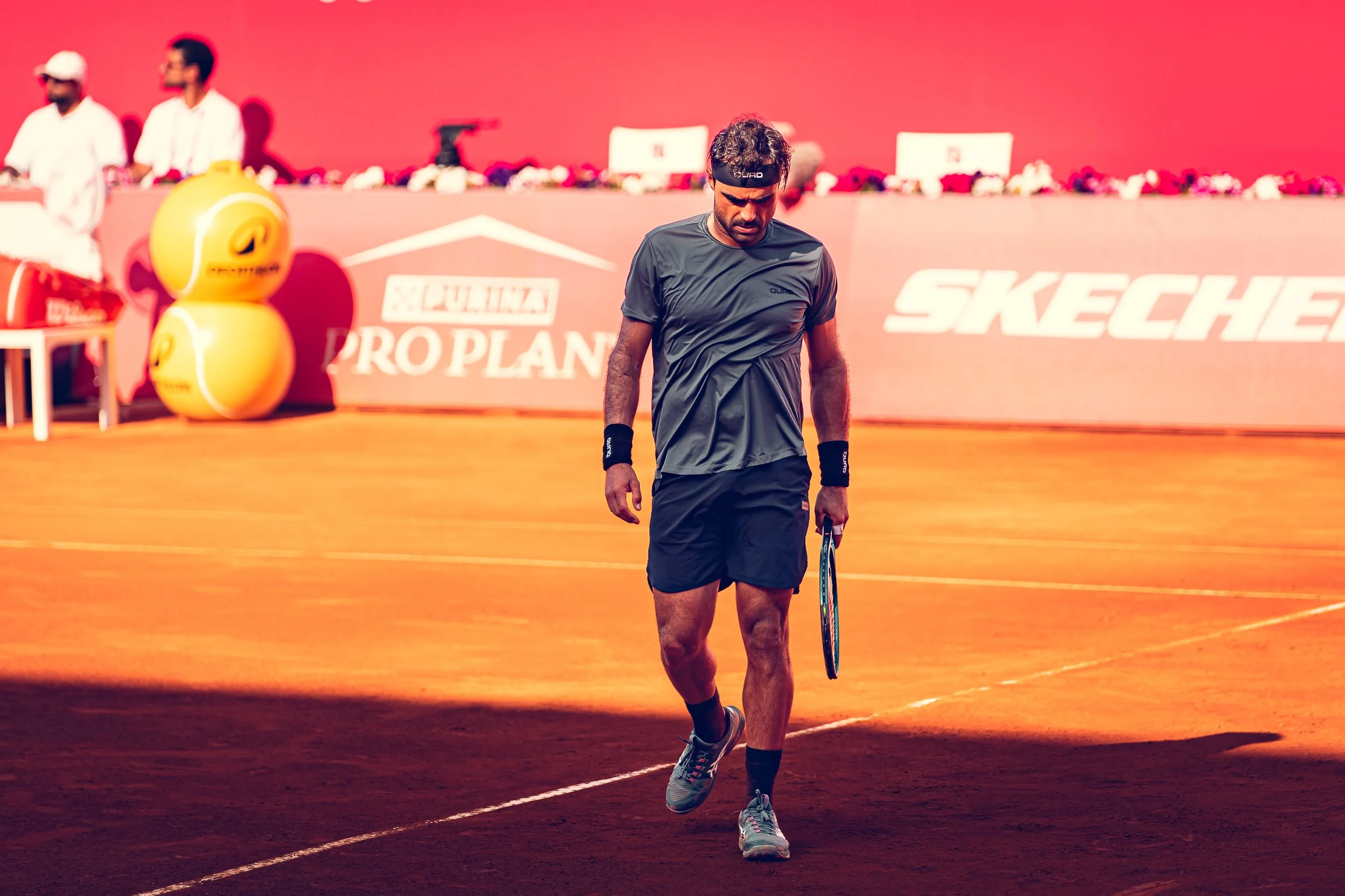A male tennis player walking on a clay court with a tennis racket, wearing black sportswear and a headband, during a match or practice session.