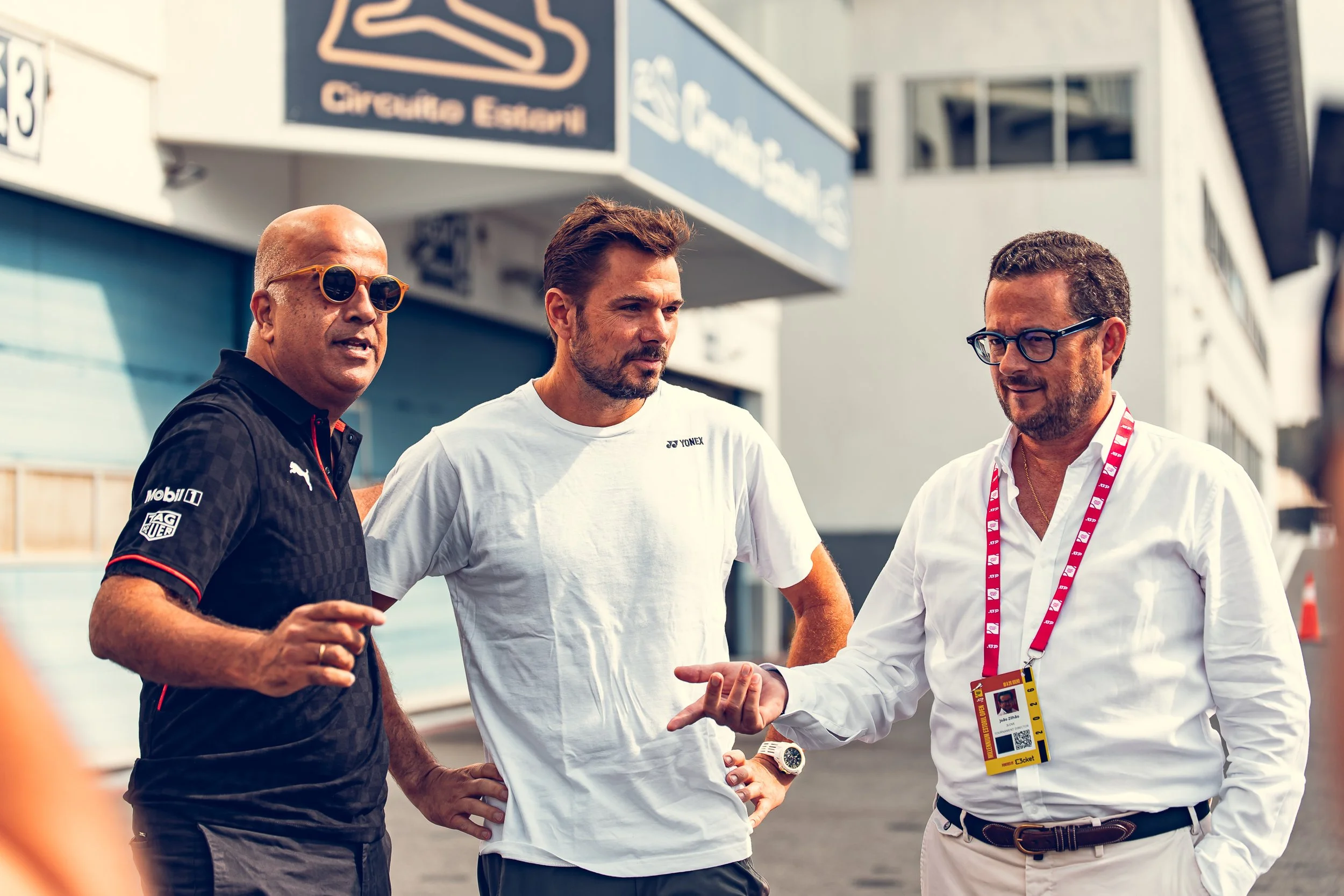 Three men standing outdoors, engaged in conversation. The man on the left is bald, wearing sunglasses, and dressed in a black shirt with sponsor logos. The man in the center has brown hair, a beard, and is wearing a white T-shirt. The man on the righ