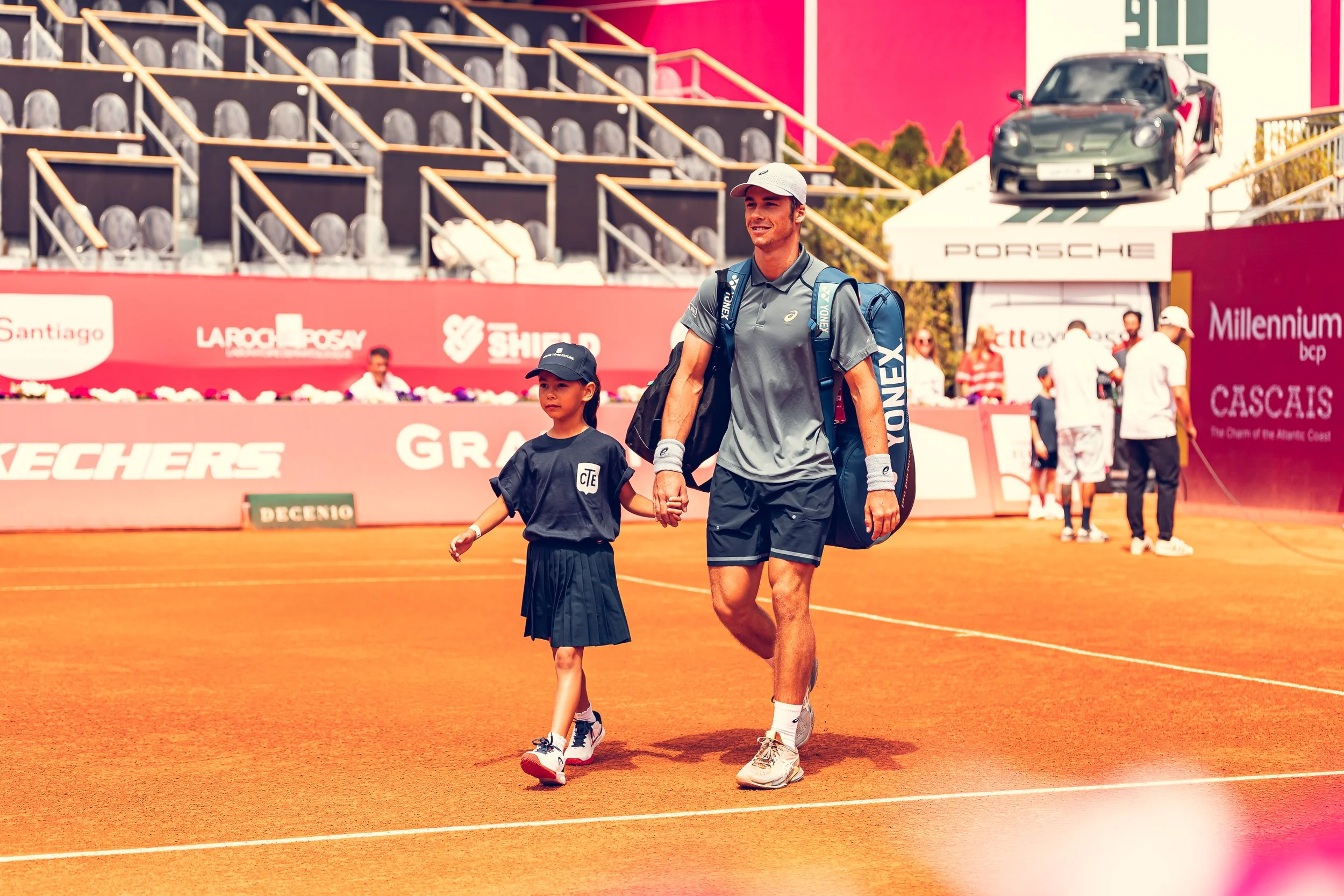 A male tennis player with a backpack walking hand-in-hand with a young girl on a clay tennis court. Behind them is a spectator stand and a hill of spectators, with a Porsche car display on a ramp. The scene appears to be at a tennis tournament.
