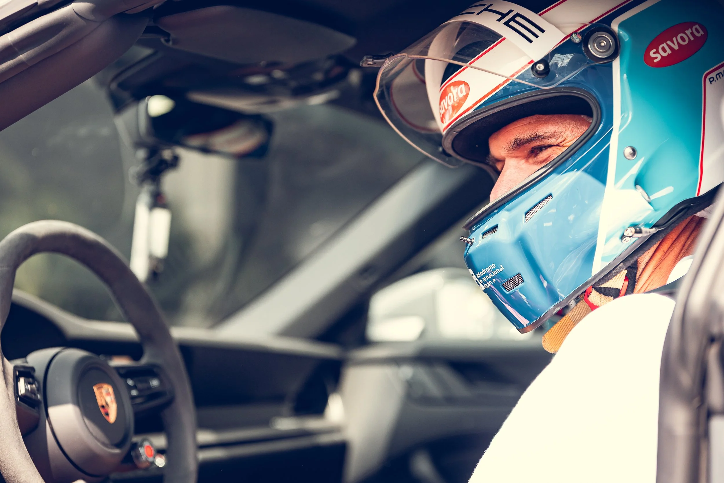 Close-up of a race car driver wearing a blue helmet with sponsor logos and looking out while seated in the cockpit of a Porsche race car.