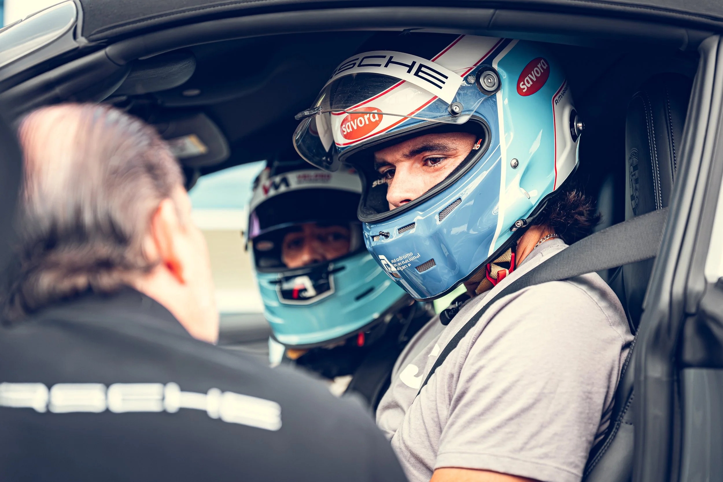 Race car driver wearing a light blue helmet with red and black graphics, sitting inside a race car and looking at someone outside the vehicle.
